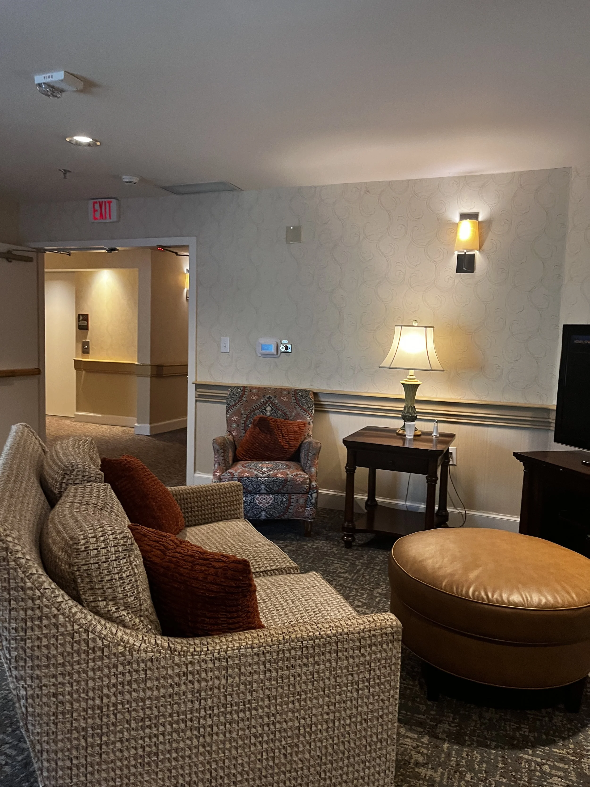 Living room with patterned sofa, orange pillows, an armchair with a floral pattern, a side table with a lamp, and a wall-mounted light, in a hotel or hotel-like setting.