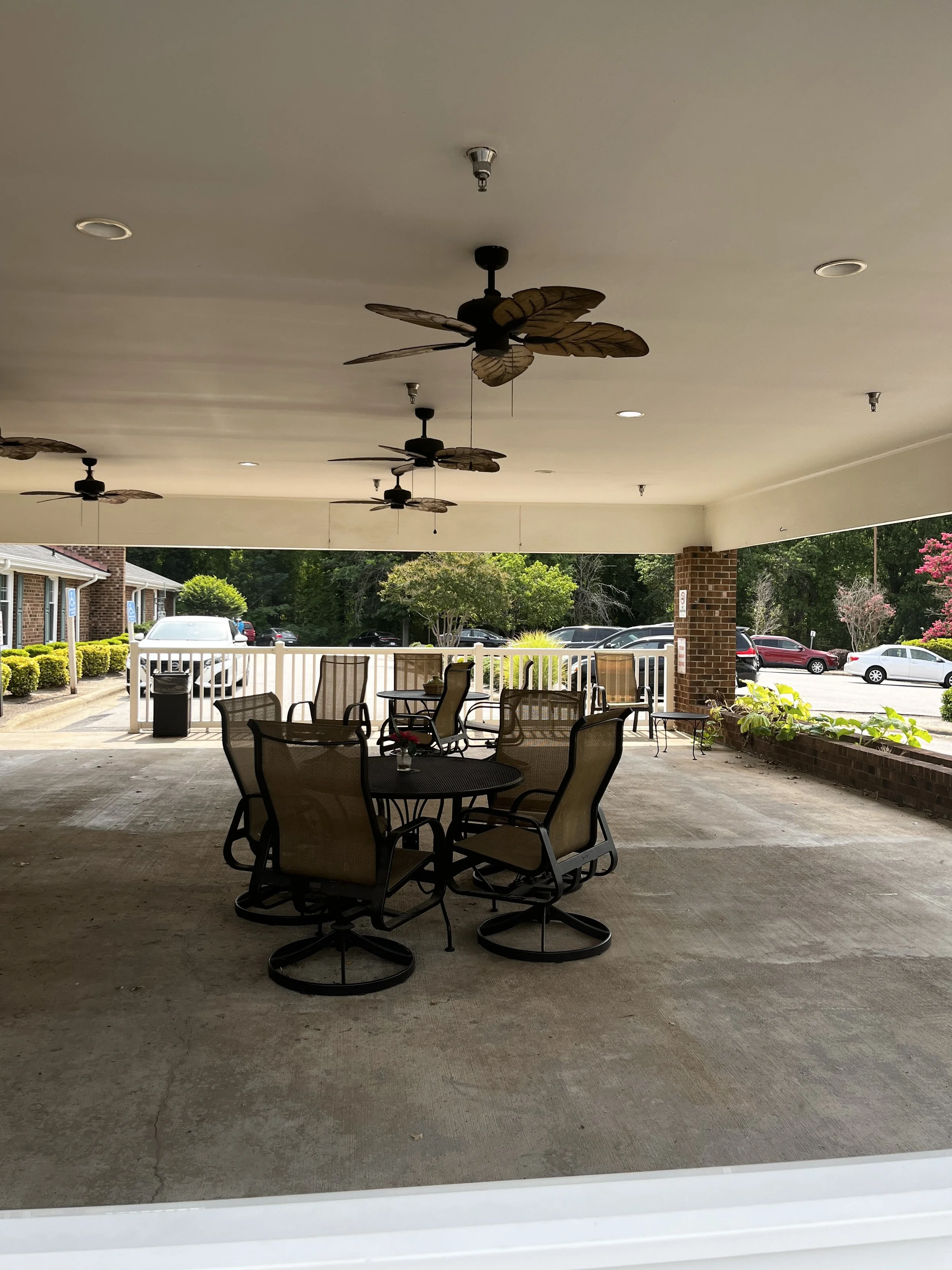 View of an outdoor patio with round tables and chairs, ceiling fans with leaf-shaped blades, and a parking lot with cars and greenery beyond.