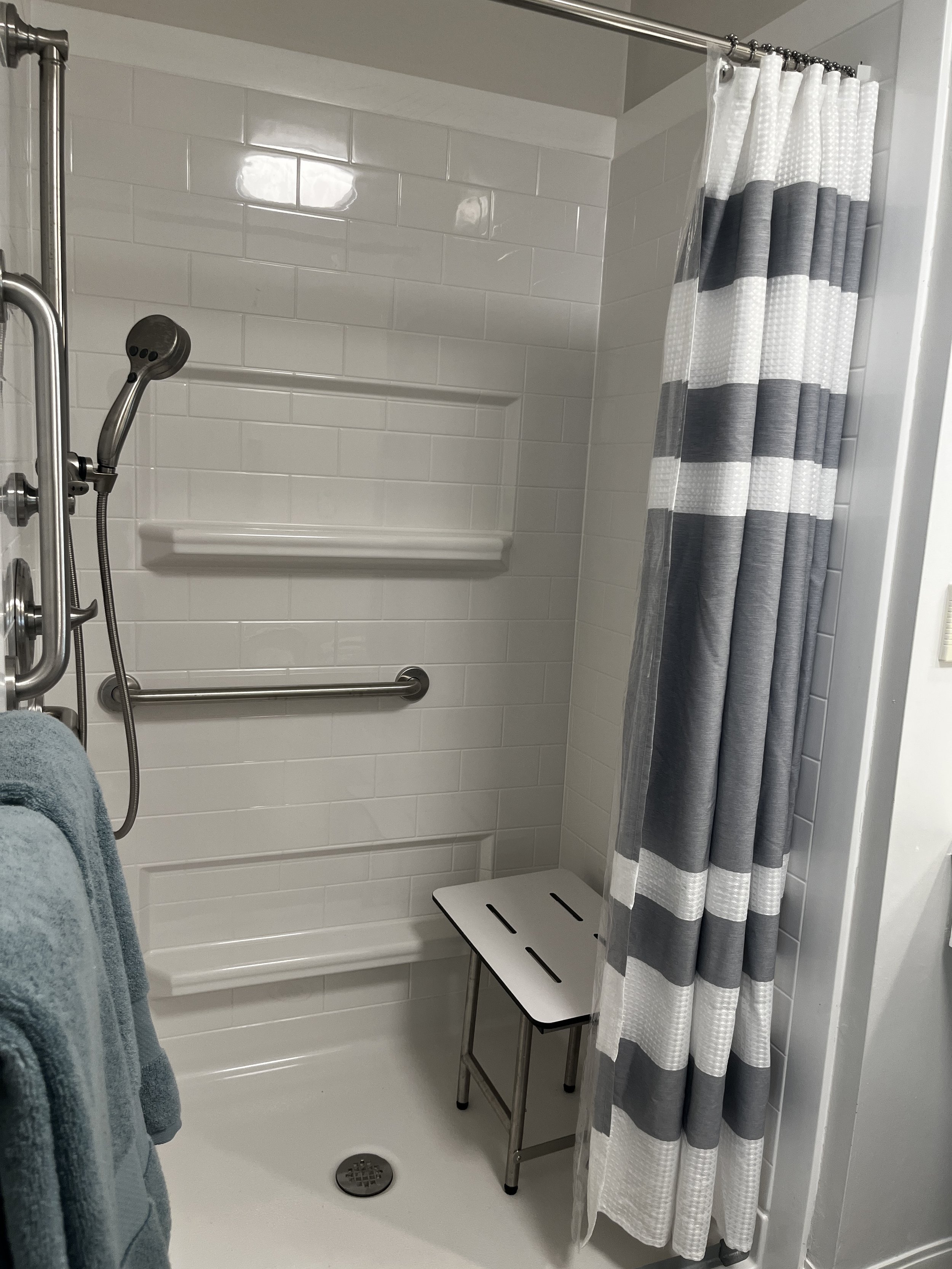 A walk-in shower with white subway tile walls, a stainless steel adjustable shower head, a shower seat, a silver grab bar, a striped shower curtain in shades of gray and white, and a small black metal table inside the shower area.