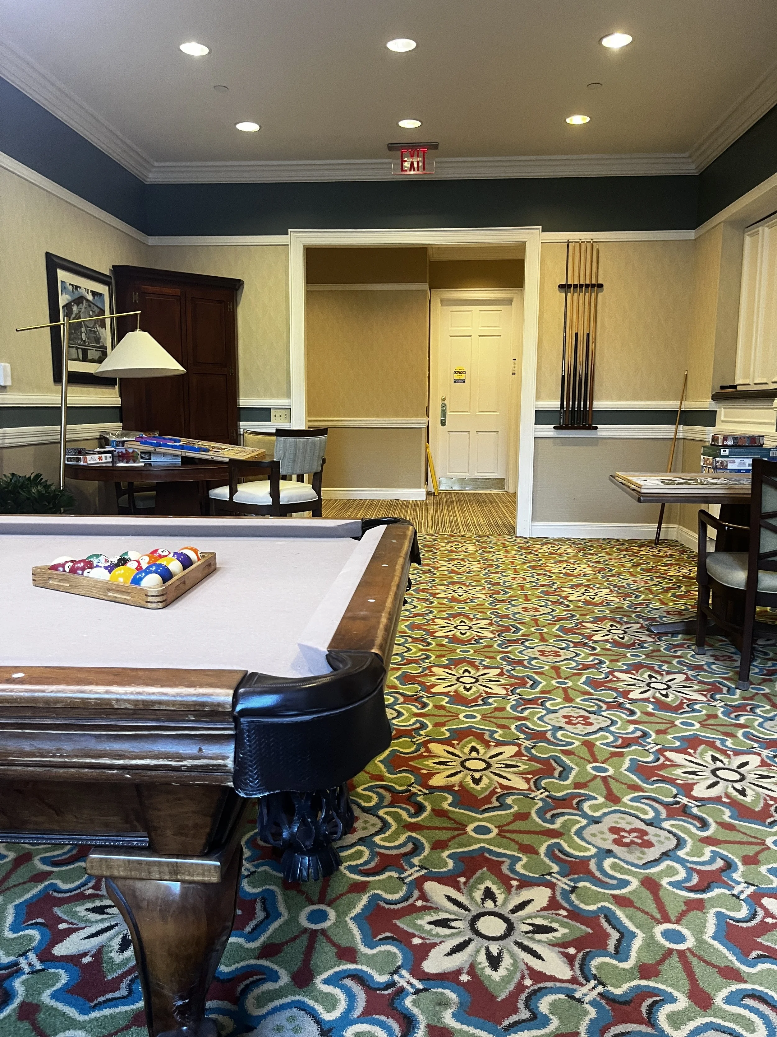 A room with a billiards table in the foreground, a colorful patterned carpet, a table with board games, a wall-mounted cue stick rack, and a door in the background.