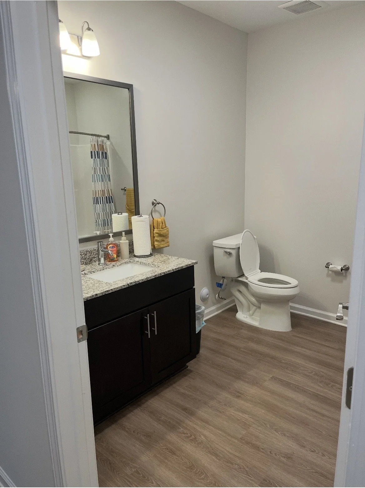 A bathroom with a vanity with a granite countertop, soap dispenser, paper towels, and a mirror. A toilet is in the corner next to a toilet paper holder. The floor is wooden, and the walls are plain white.