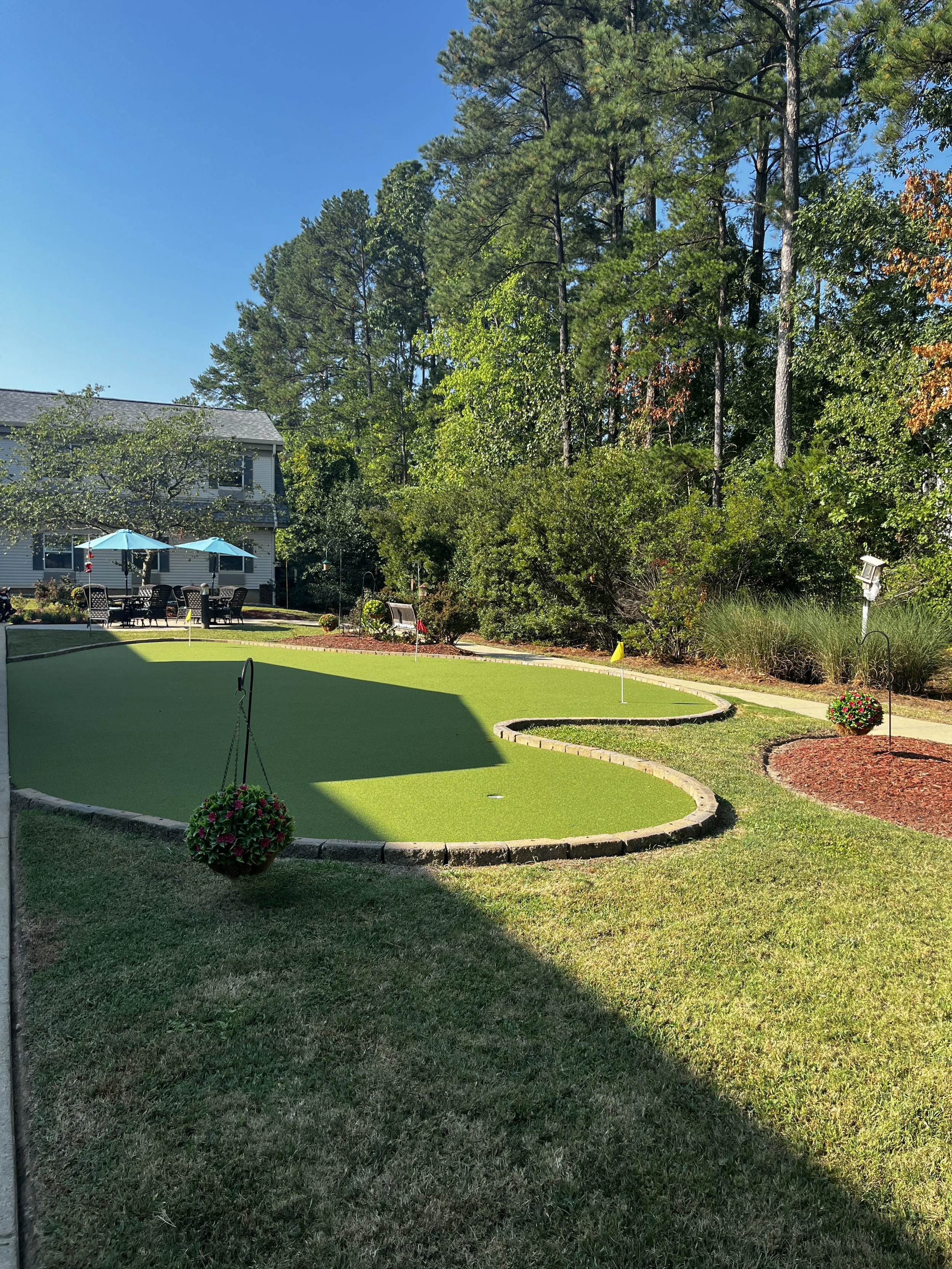 A backyard putting green with a flag in the center, surrounded by grass, brick edging, hanging flower baskets, and outdoor furniture with umbrellas, adjacent to a house and tall trees in the background.