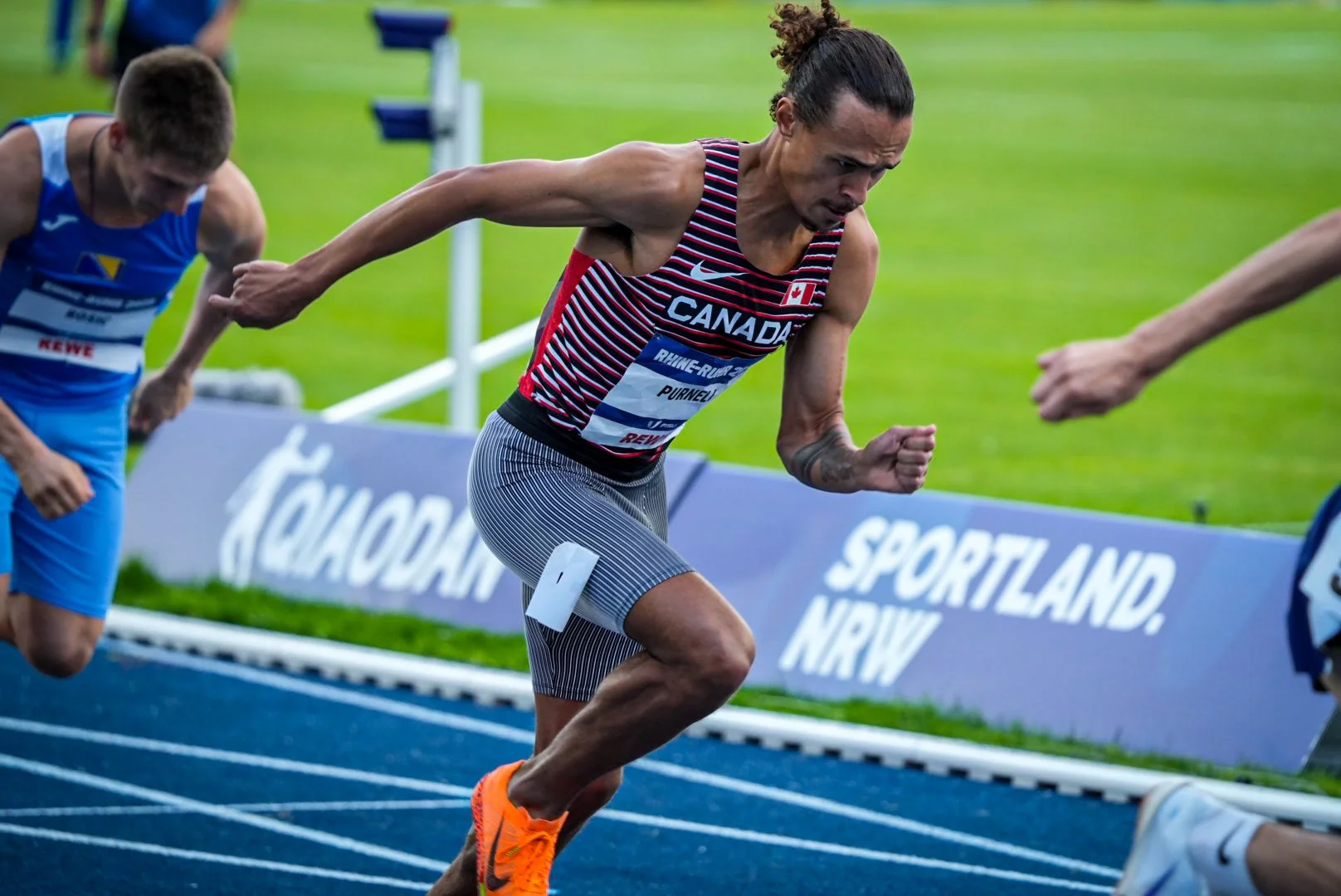 A man competing in a race, wearing a Canada athletic uniform, is sprinting on a blue track with an intense focus. Other runners and a sign with 'SPORTLAND, NRW' are visible in the background.