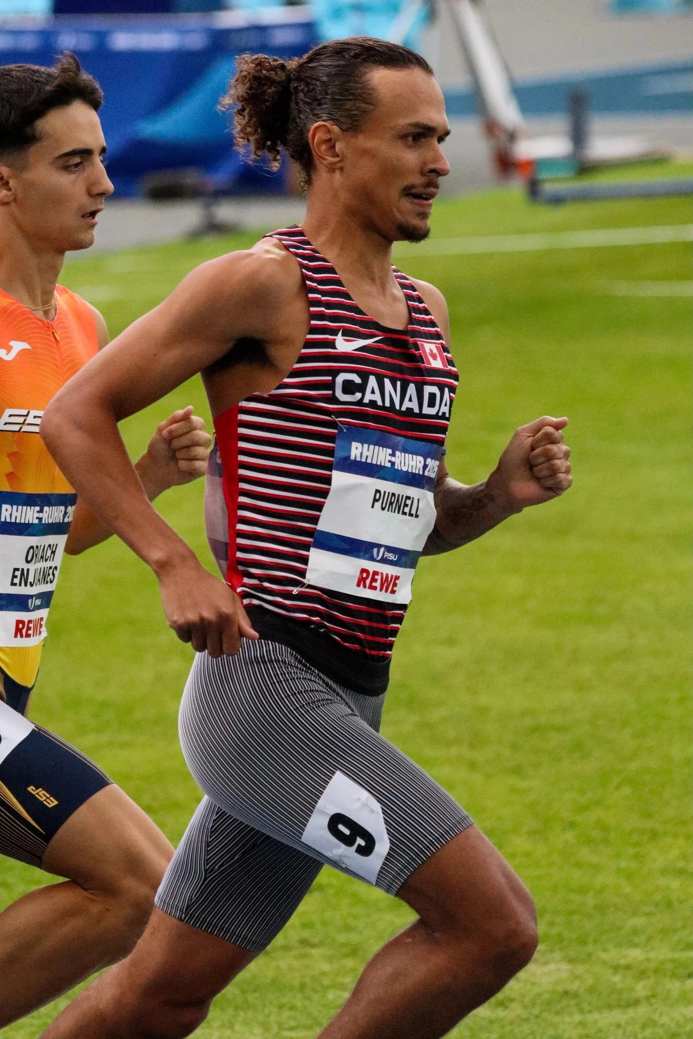 Two male athletes running on a grassy track during a race, with one athlete in a black and white striped uniform and the other in an orange uniform. The athlete in the foreground has a focused expression.