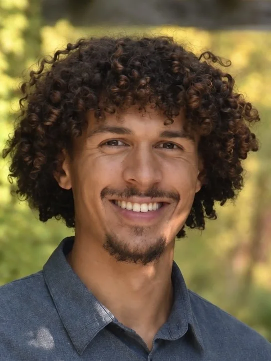A smiling man with curly dark hair and a goatee, wearing a denim shirt, outdoors with blurred greenery in the background.