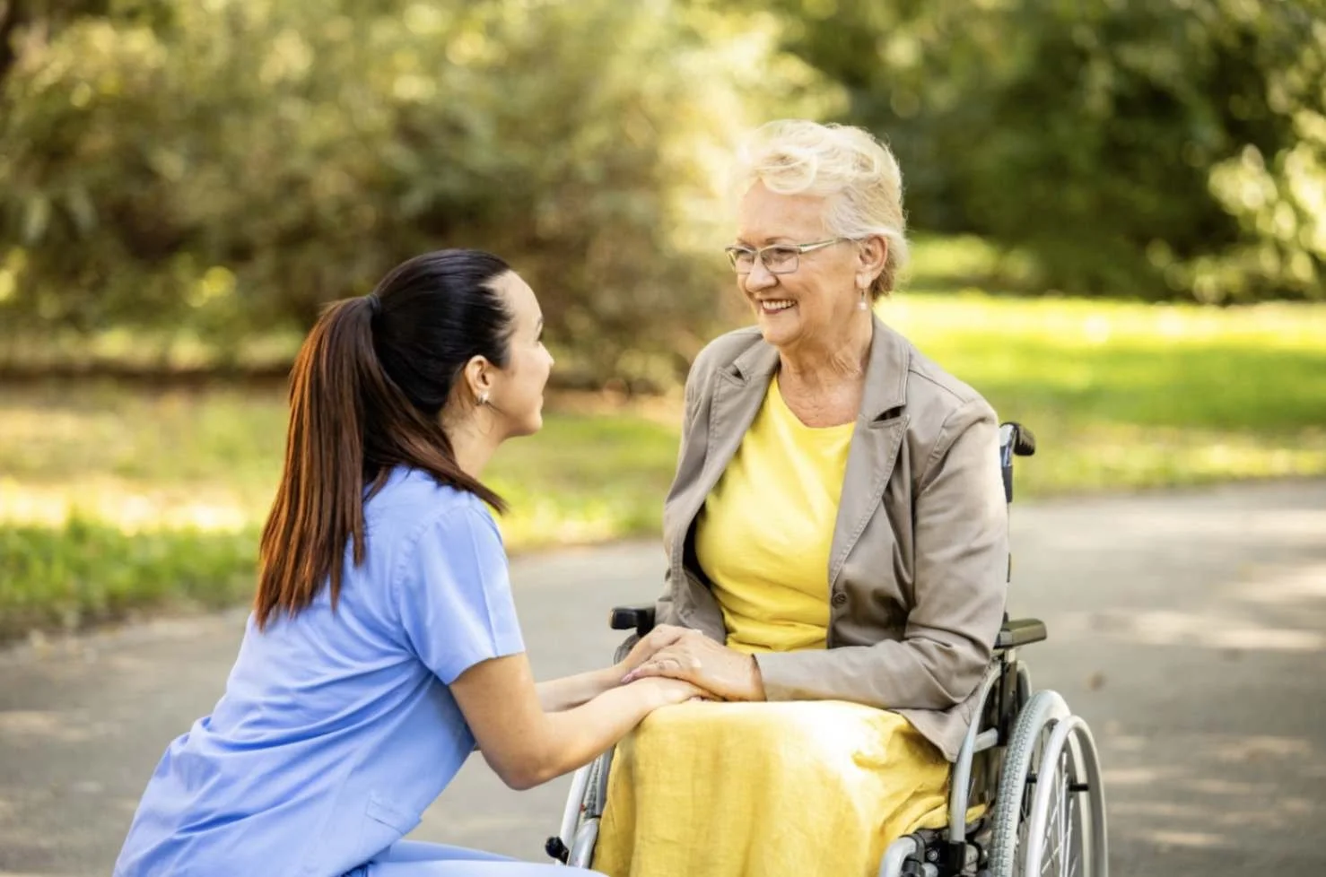 An assisted living caregiver in blue scrubs holding hands and smiling with a senior woman in a wheelchair outdoors in a park.