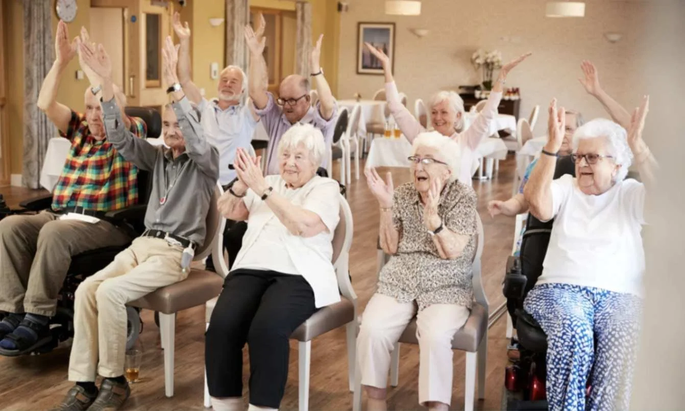 Seniors sitting in a group class raising their hands and clapping during a wellness activity at an assisted living facility.