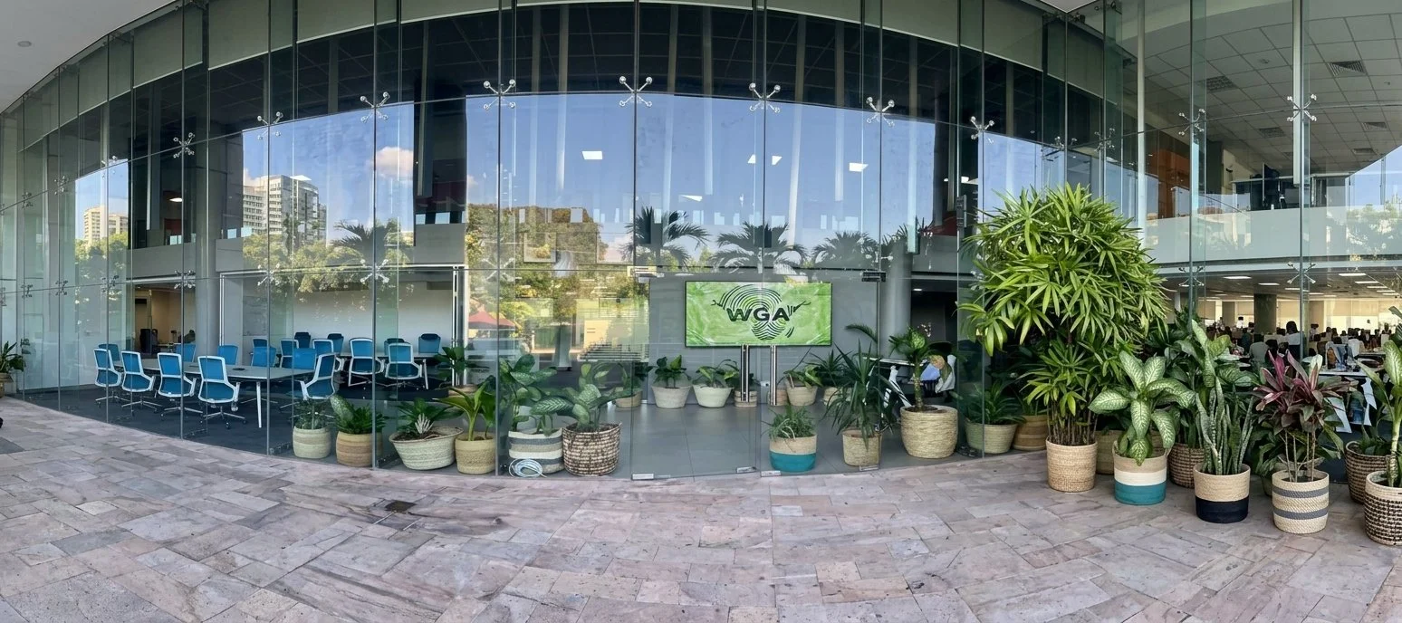 Exterior view of a glass building with plants outside, and an indoor office with desks and people visible through the glass.
