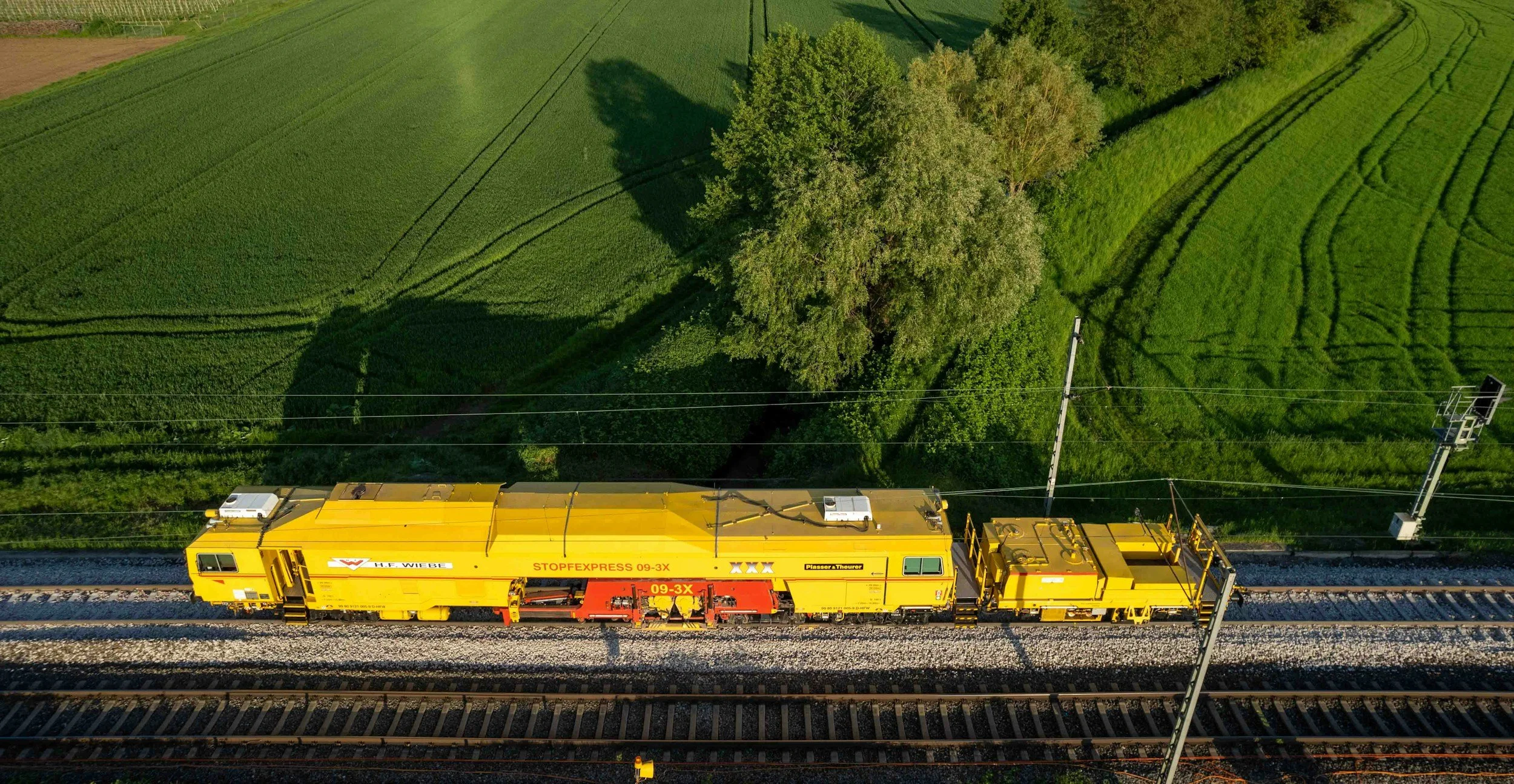 A yellow maintenance train running on railway tracks surrounded by green fields and trees.
