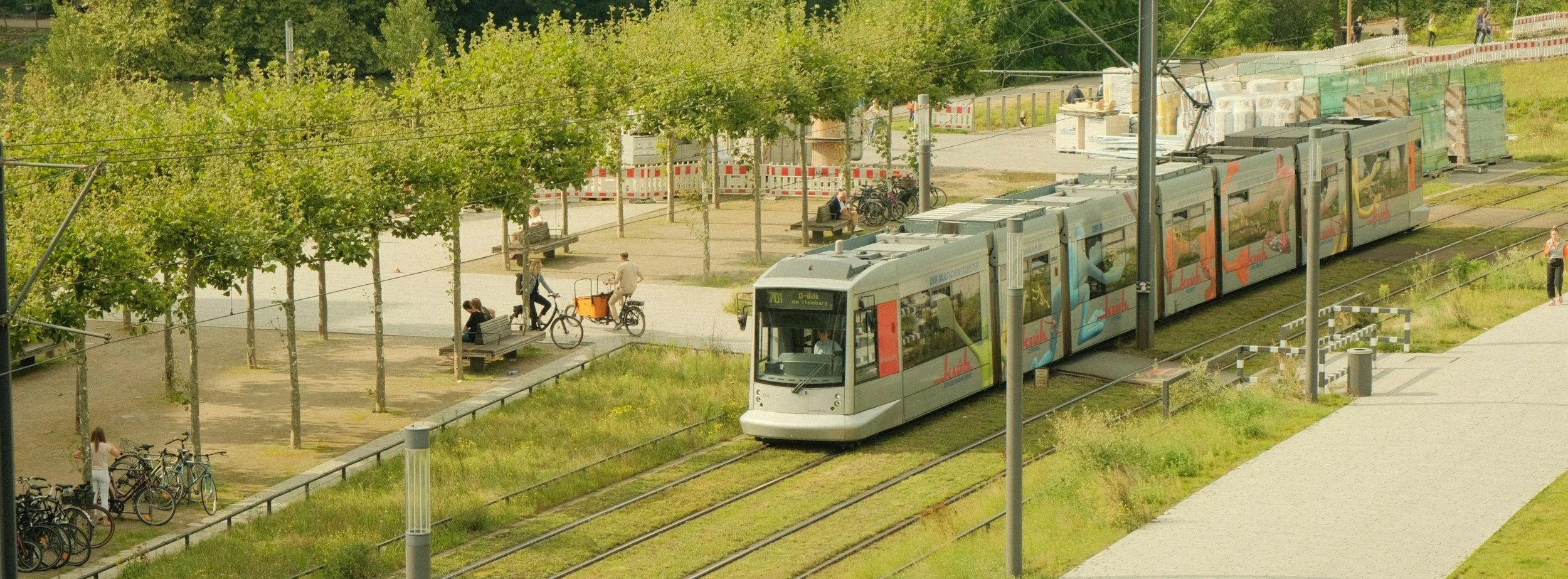 A light rail train traveling through a park area with trees, benches, and people walking and riding bikes along the sidewalk.