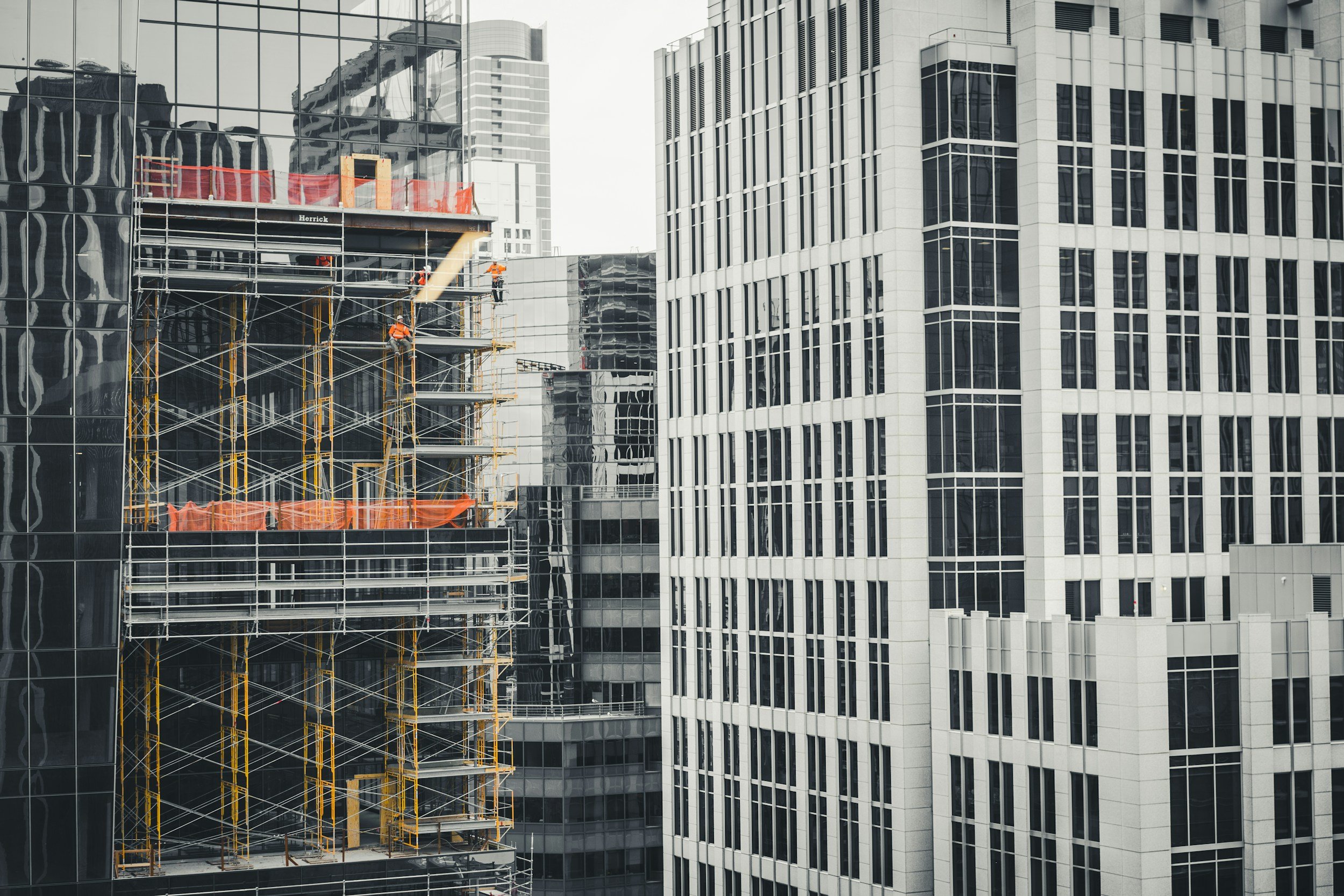 Construction workers working on scaffolding on a high-rise building in an urban area, with other tall modern glass buildings surrounding them.