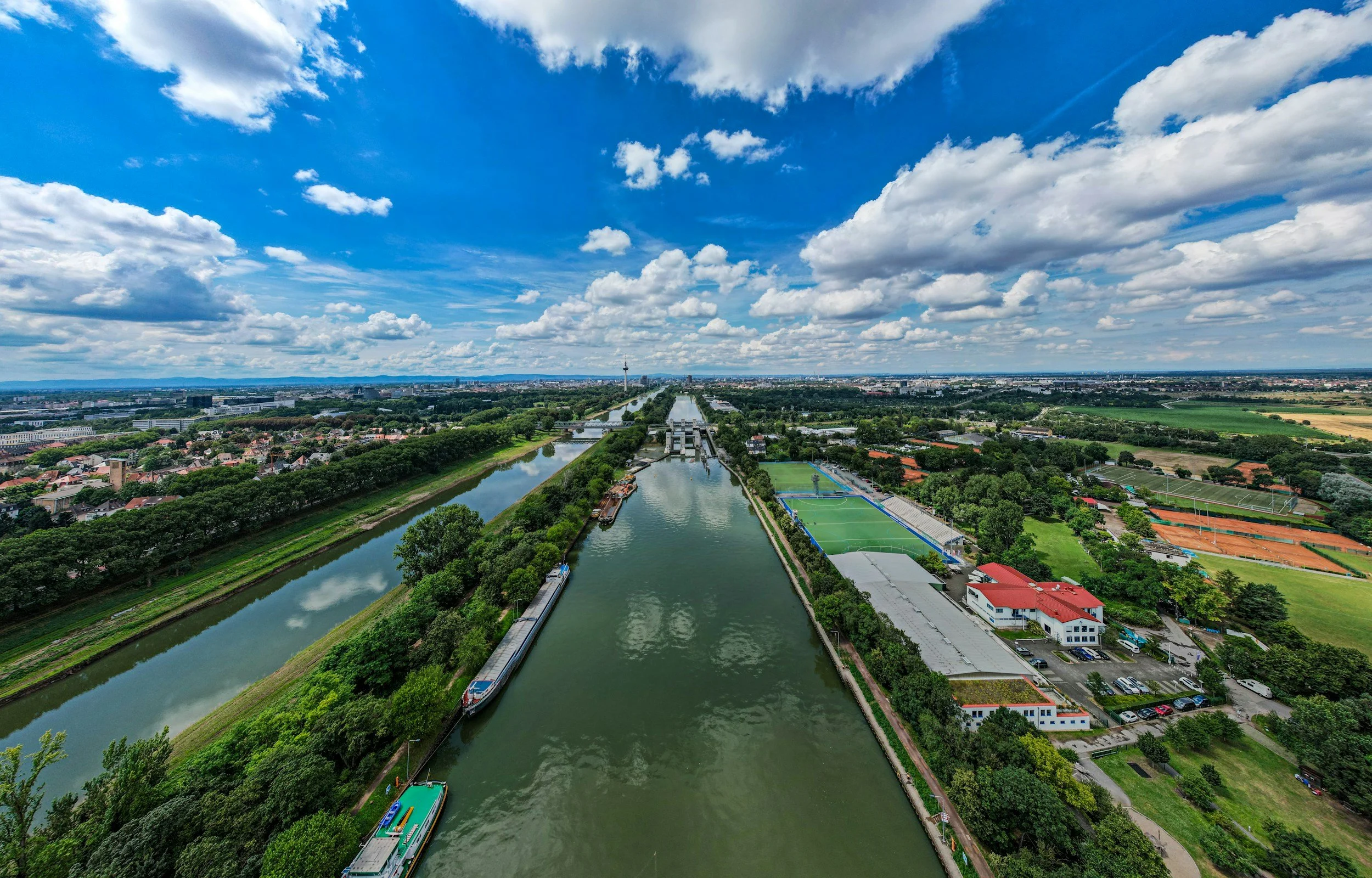 A panoramic view of a city with a river running through it, featuring a boat, green parks, residential houses, sports fields, and a tower in the distance under a partly cloudy sky.