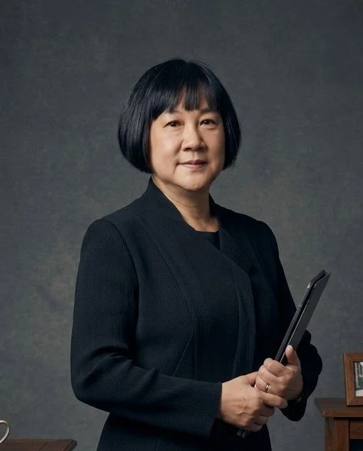 Woman with short black hair in a black suit holding a tablet, standing in front of a gray background