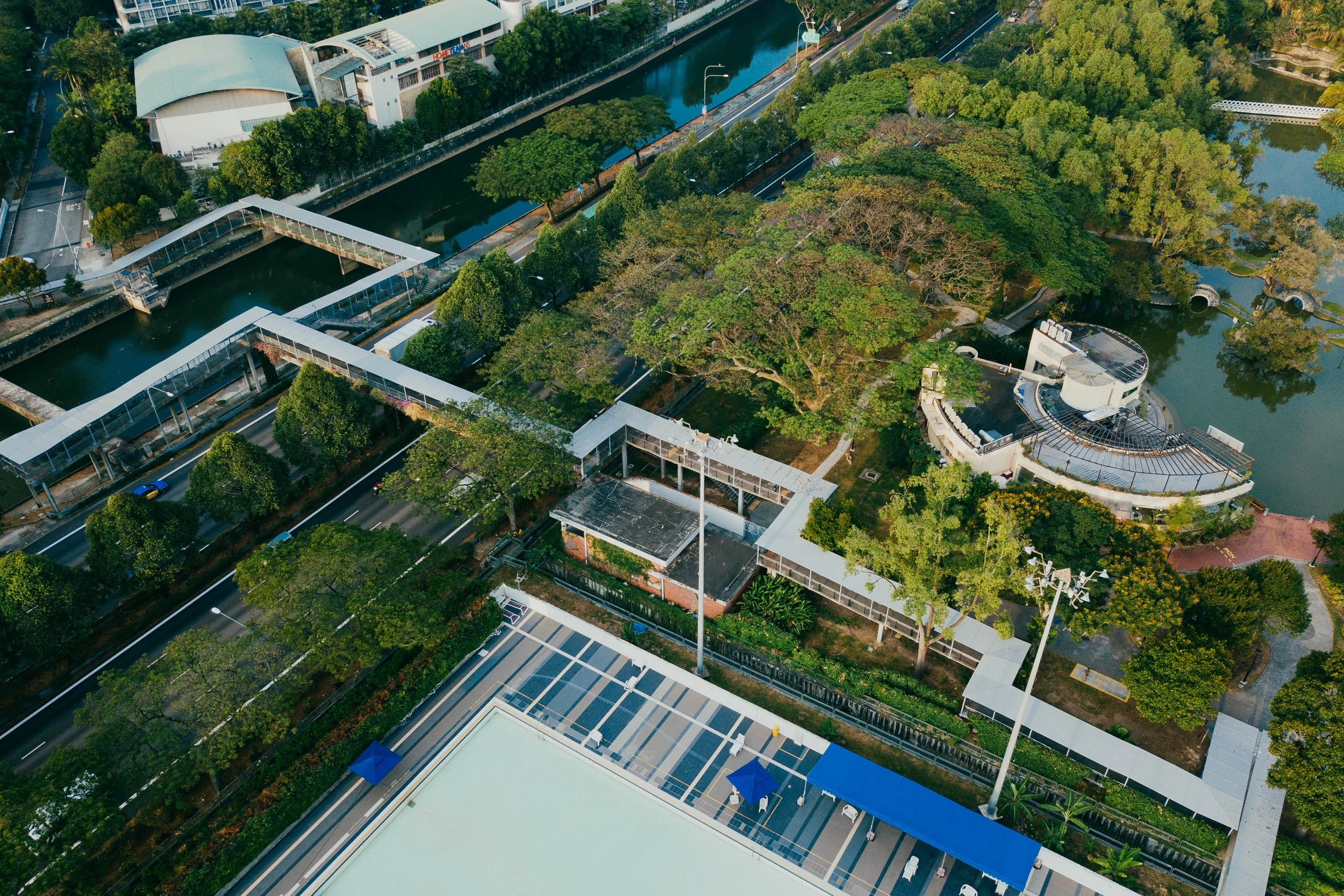 An aerial view of a city park with trees, pathways, water features, and modern buildings surrounding the area.