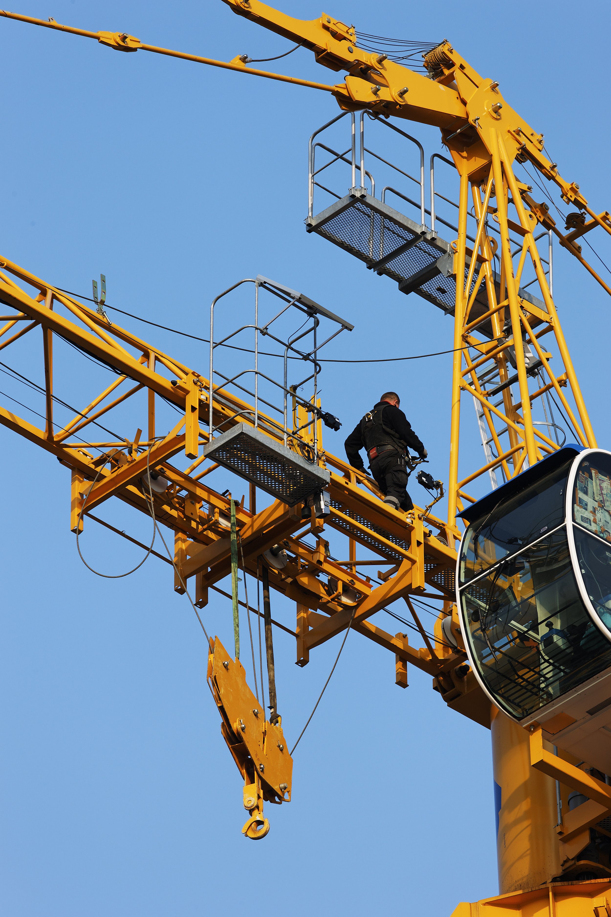 A construction worker wearing a black jacket and gray pants stands on a yellow construction crane platform against a bright blue sky.