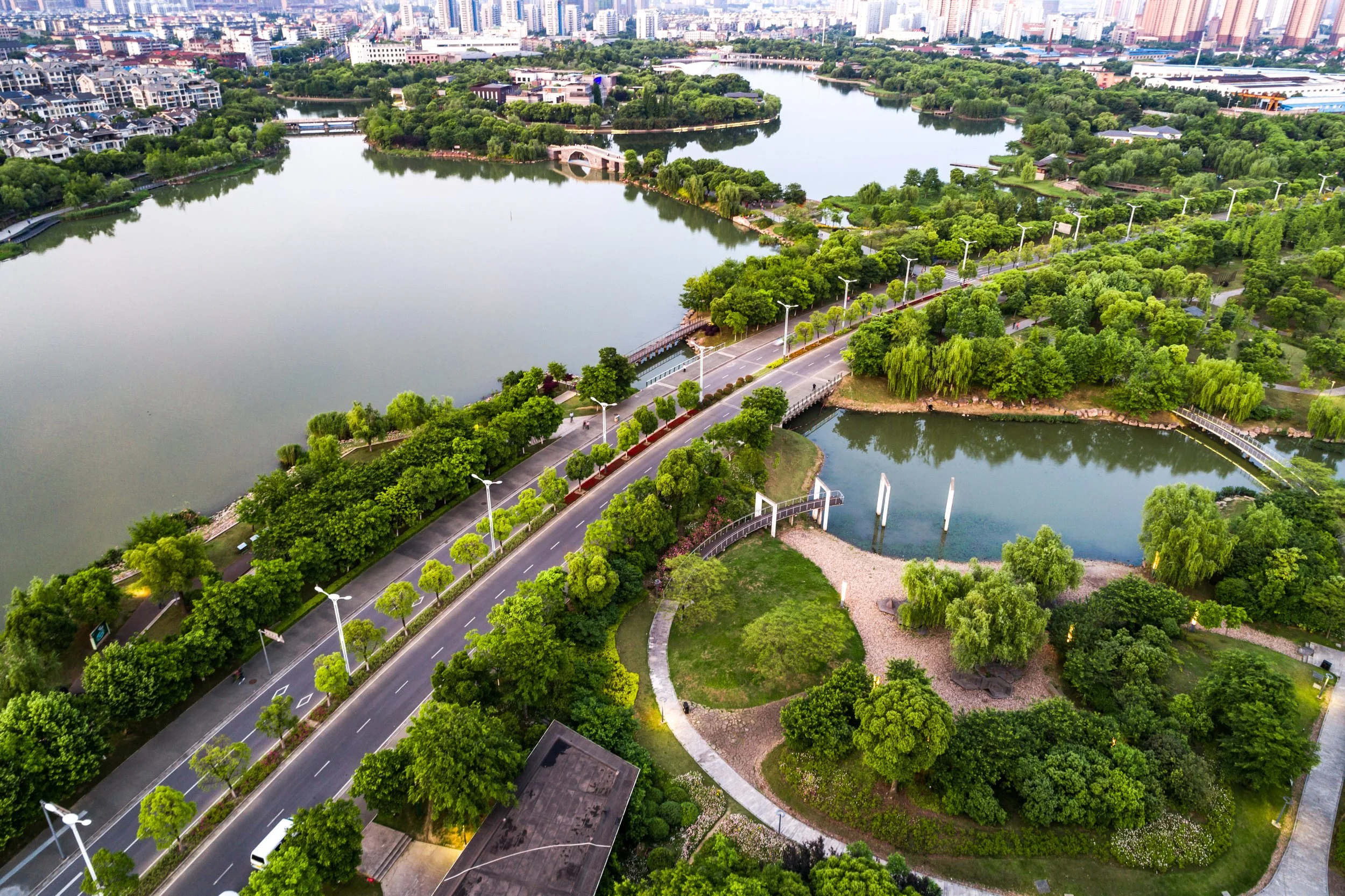 Aerial view of a city park with a lake, surrounded by trees, pathways, and bridges, with city skyscrapers in the background.