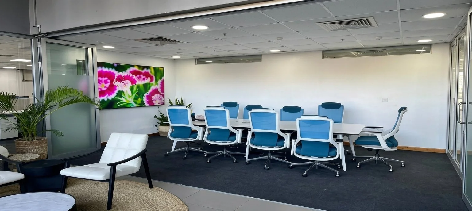 Office conference room with a large white table, blue and white office chairs, a flat-screen TV showing pink flowers, potted plants, and seating area with white chairs and a rug.