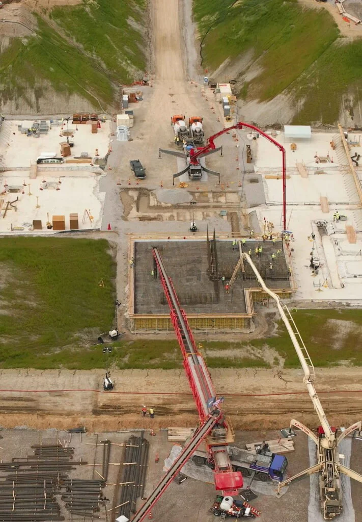 An aerial view of a construction site showing concrete pumps, trucks, and workers building a foundation for a structure with steel reinforcement bars.
