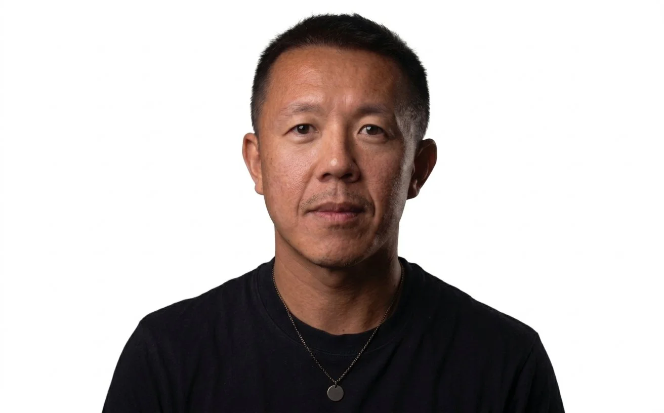 A portrait of an Asian man with short black hair, wearing a black t-shirt and a round pendant necklace, against a white background.
