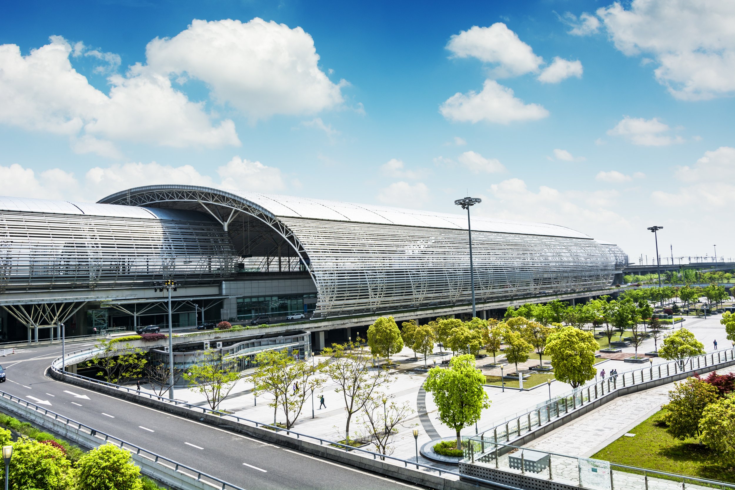 Modern transportation hub with a large, curved, metallic roof structure, adjacent parking area, landscaped greenery, and a blue sky with scattered clouds.