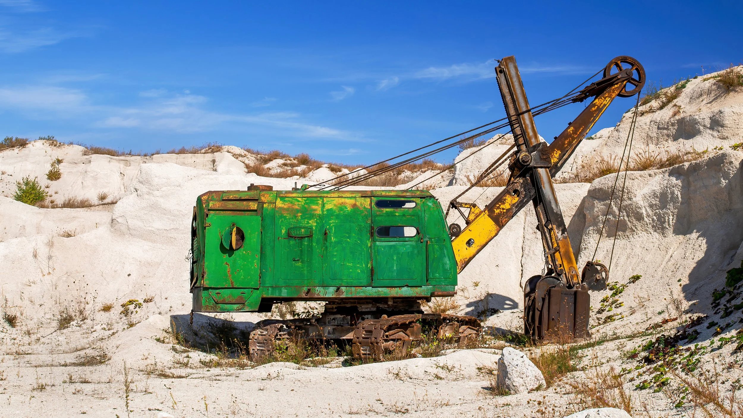 Old, green and yellow hydraulic excavator in a sandy, arid landscape with white dunes and sparse vegetation under a blue sky.