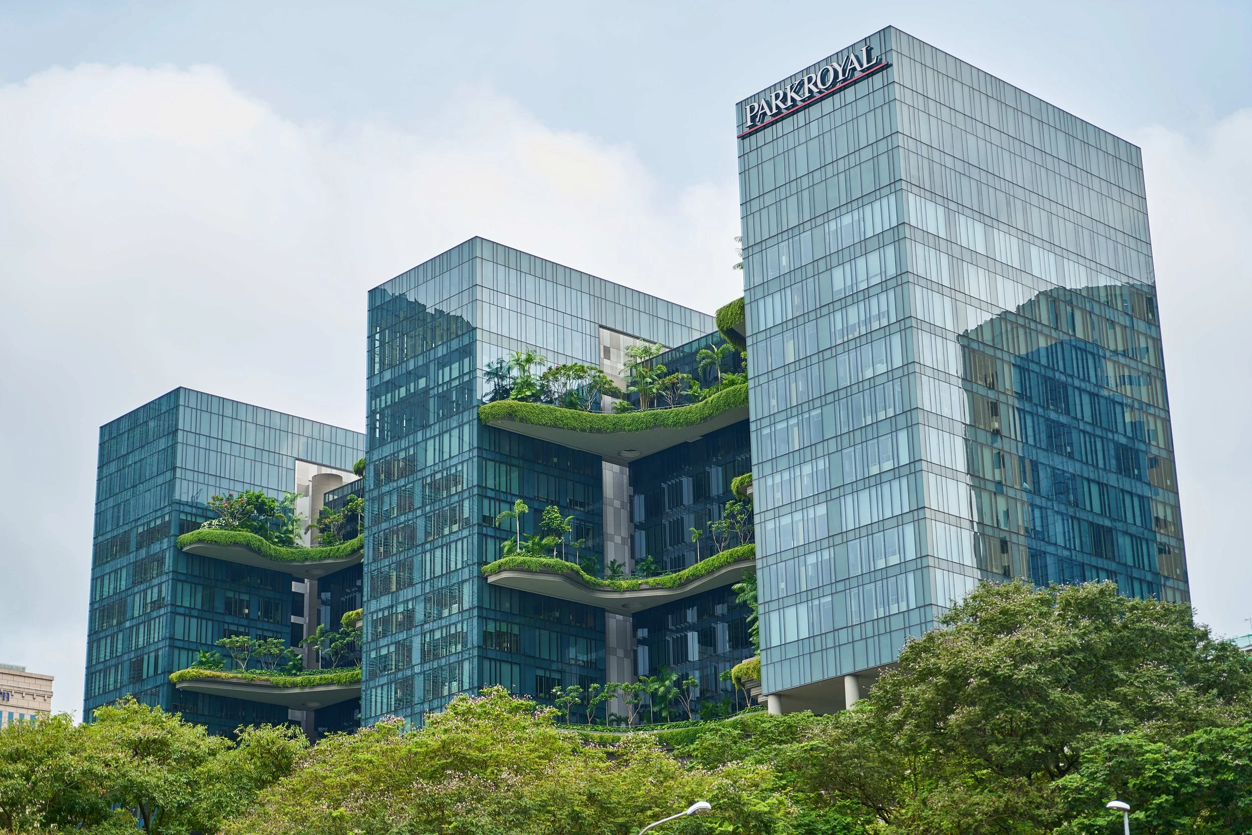 Modern glass skyscraper building with greenery on the terraces and trees in the foreground.