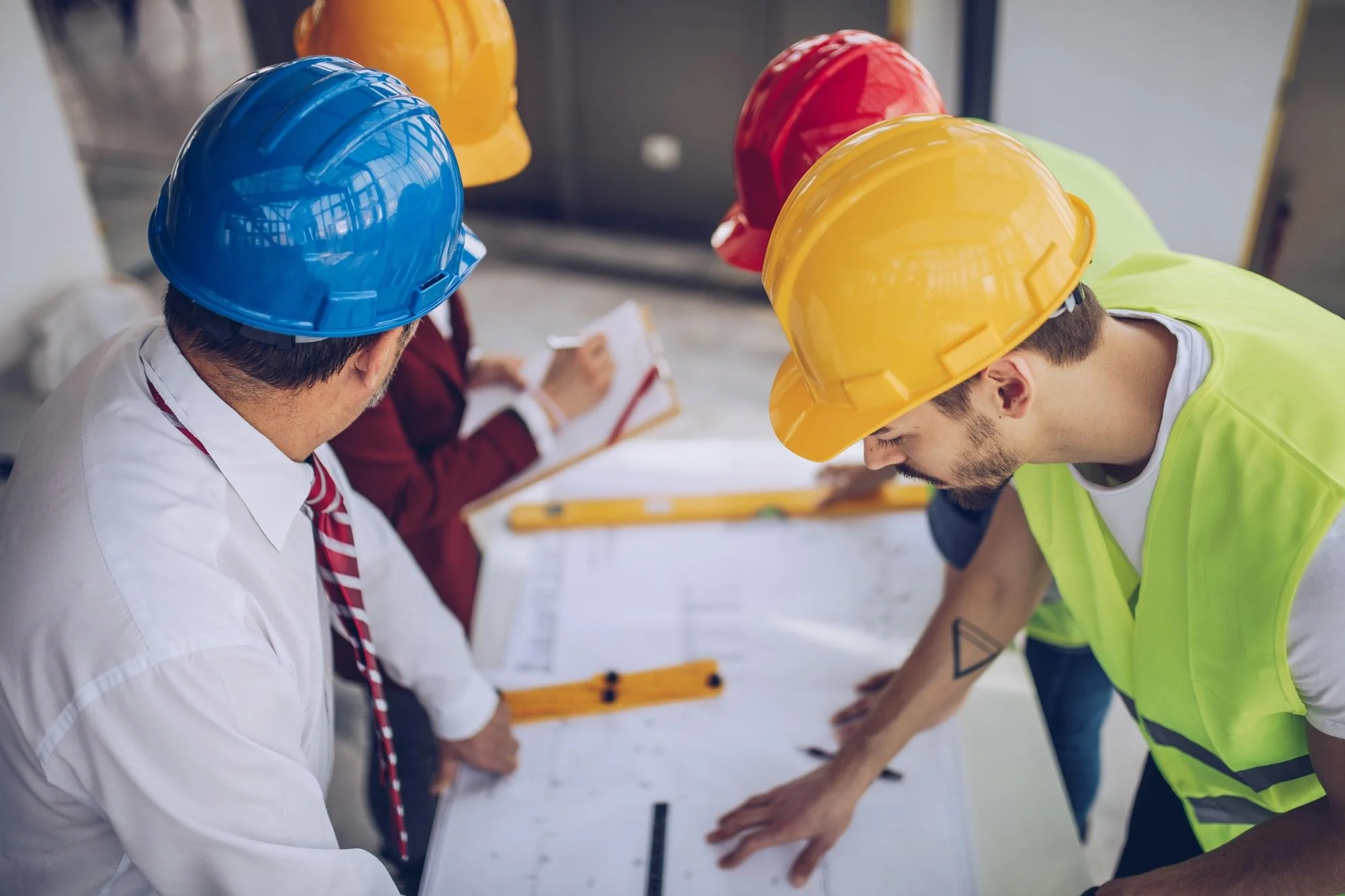 Construction workers wearing yellow, red, and blue safety helmets, working together on a structural project indoors.
