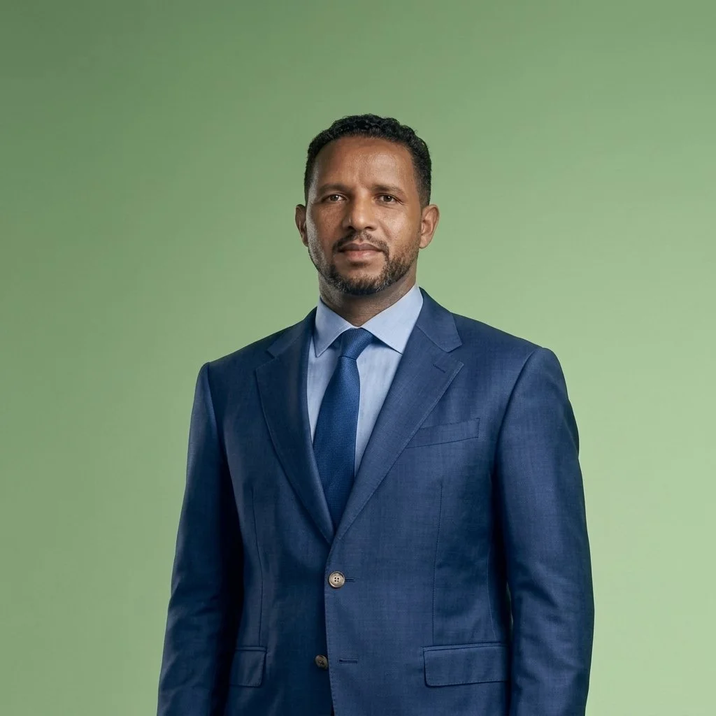 A man wearing a blue suit, light blue shirt, and matching blue tie, standing against a solid green background.