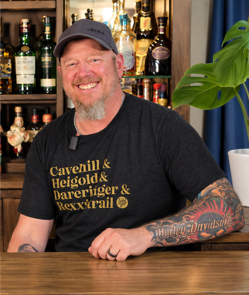Smiling bearded man in a cap and black T-shirt with tattooed arms sitting at a wooden bar surrounded by liquor bottles and a monstera plant