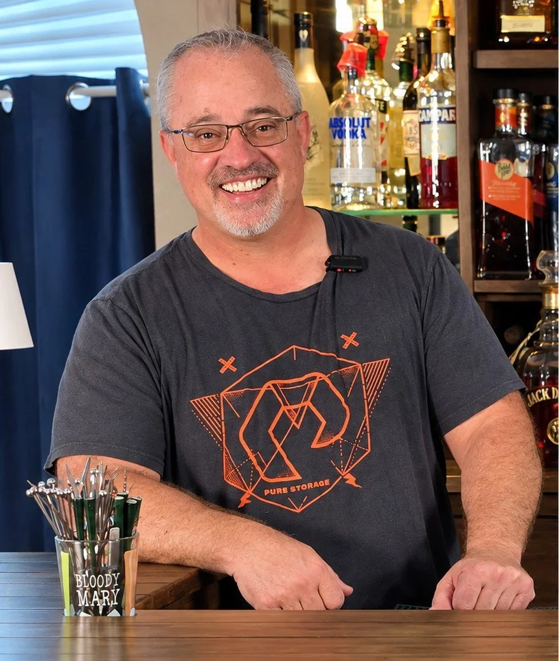 Smiling bartender wearing glasses behind a bar with liquor bottles and Bloody Mary tools