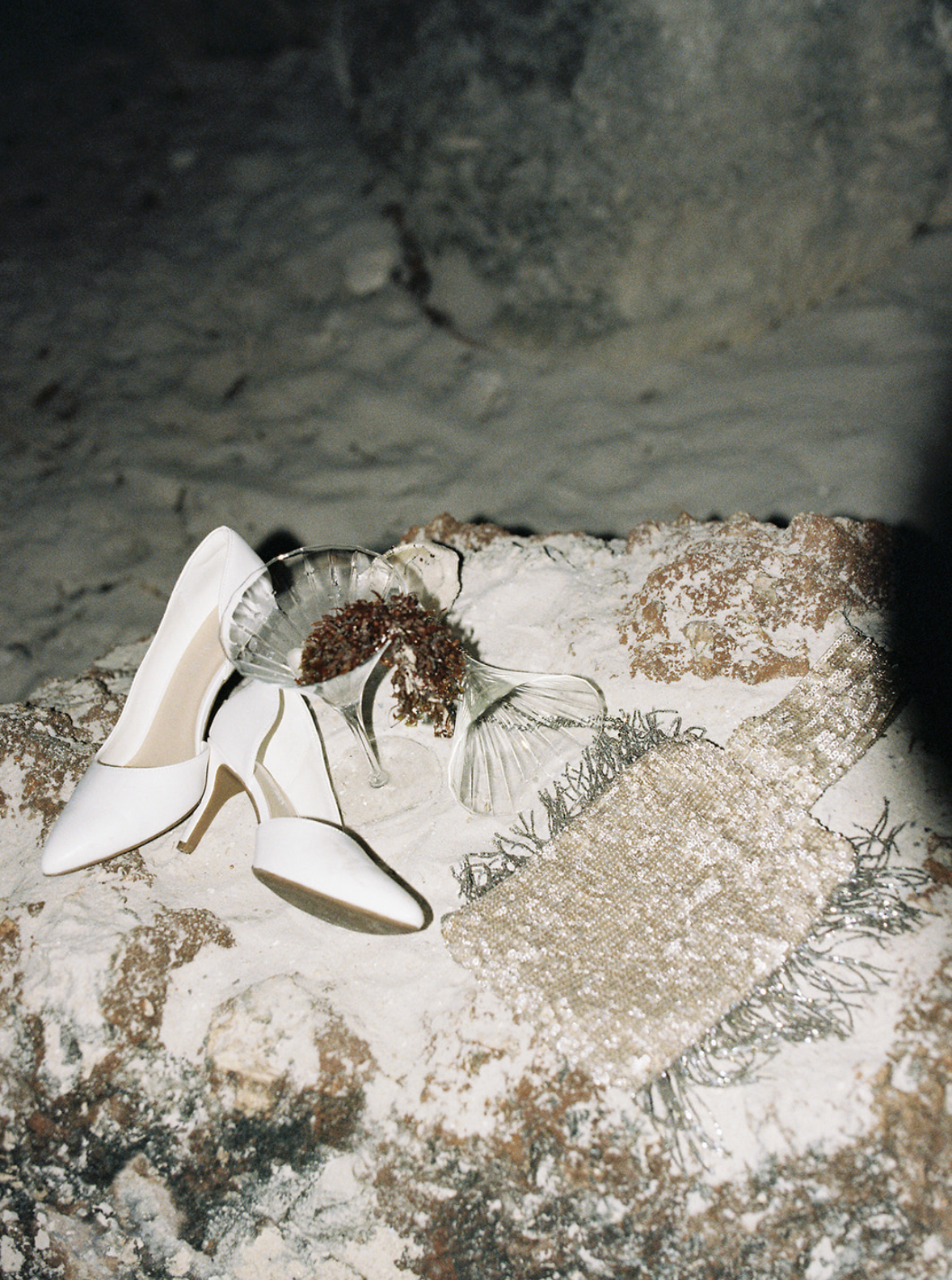 A pair of white high-heeled shoes, a glass seashell-shaped dish with dried flowers, and a woven clutch bag with metallic sequins are arranged on a sandy surface near rocks, with a dark background.