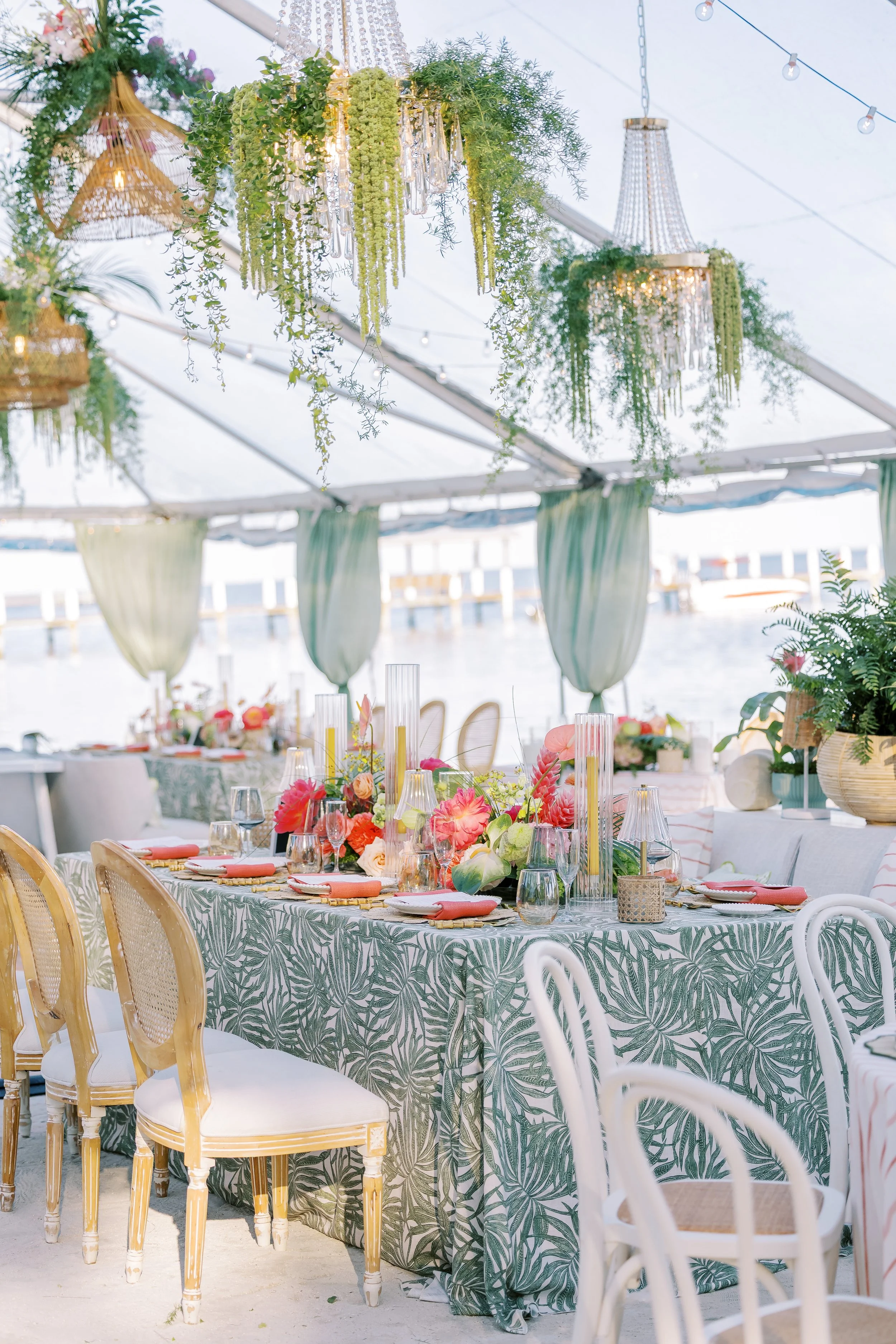 A decorated event space with a tropical-themed tablecloth, floral centerpieces, and hanging greenery and chandeliers under a tent overlooking water. Bright, summery atmosphere.