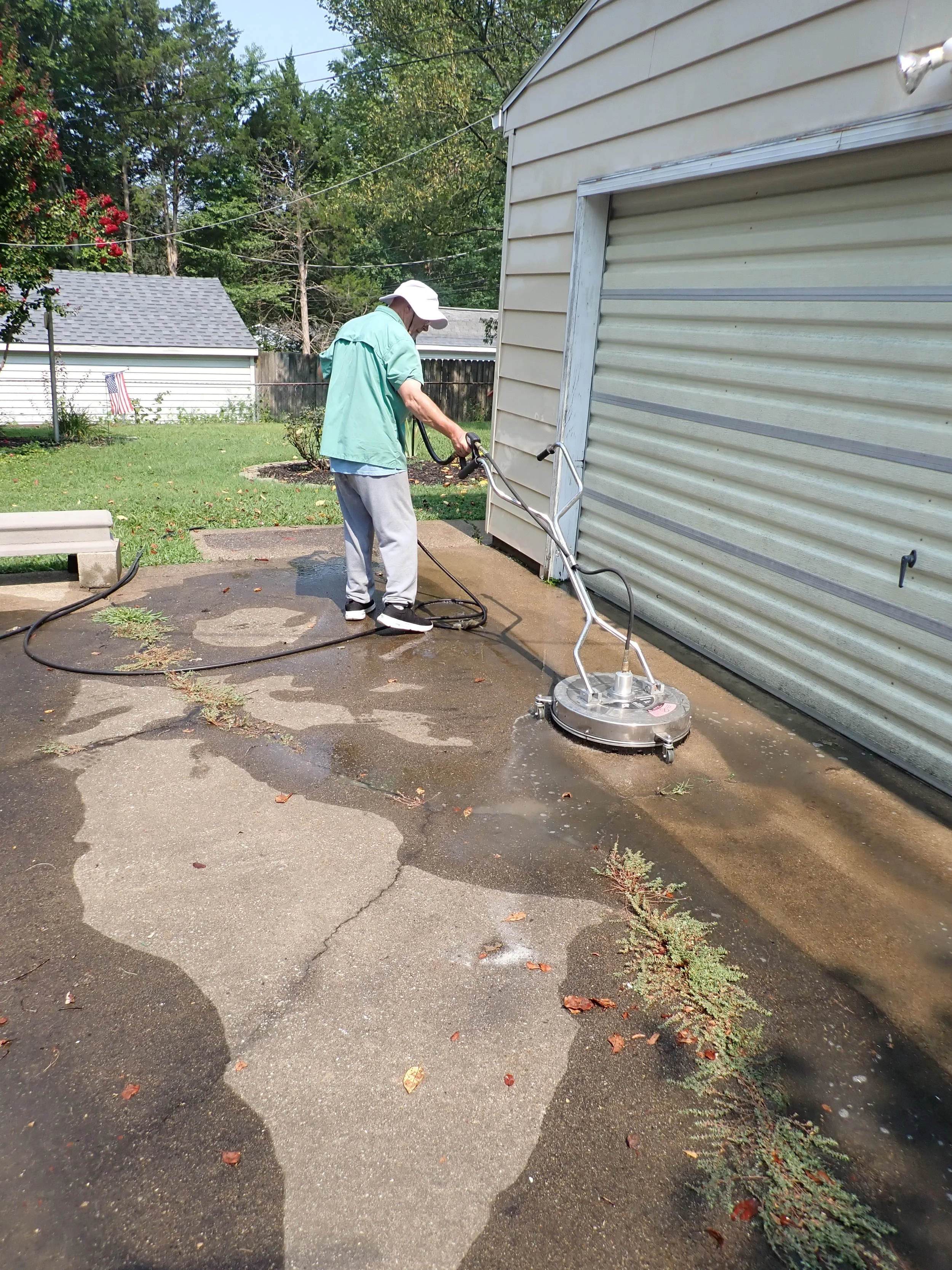 A person pressure washing a concrete driveway outside a house on a sunny day. The individual is wearing a white hat, green shirt, and gray pants, holding a pressure washer with a long hose. There are trees, a small garden, and a neighboring house in the background.