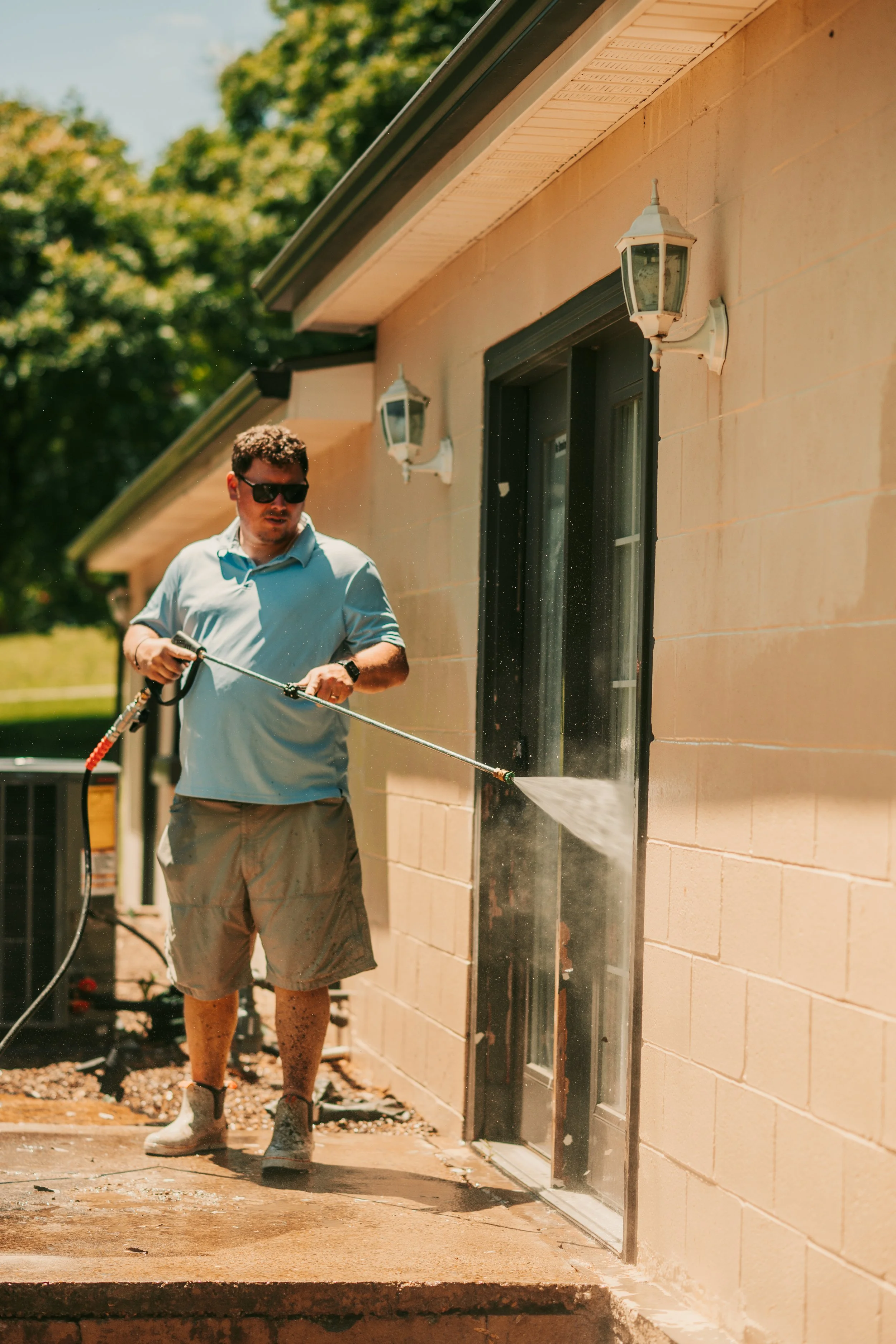 A man wearing sunglasses, a light blue shirt, beige shorts, and boots is pressure washing the exterior wall of a house. The house has beige brick walls and two outdoor wall-mounted lanterns. The background shows trees and a sunny day.