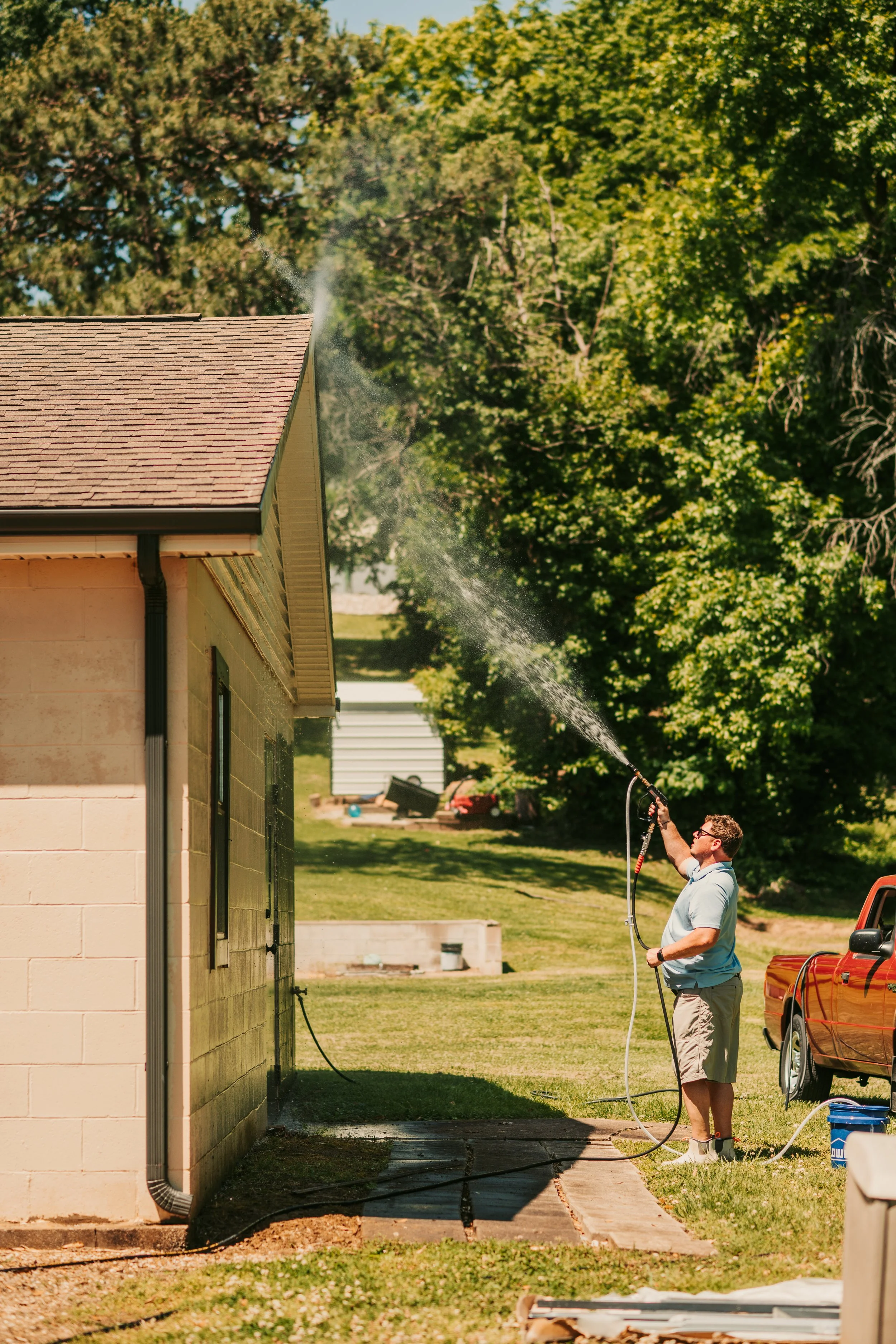 A man is watering the side of a house with a hose during daytime. The house has a brick and siding exterior, and there are trees in the background.