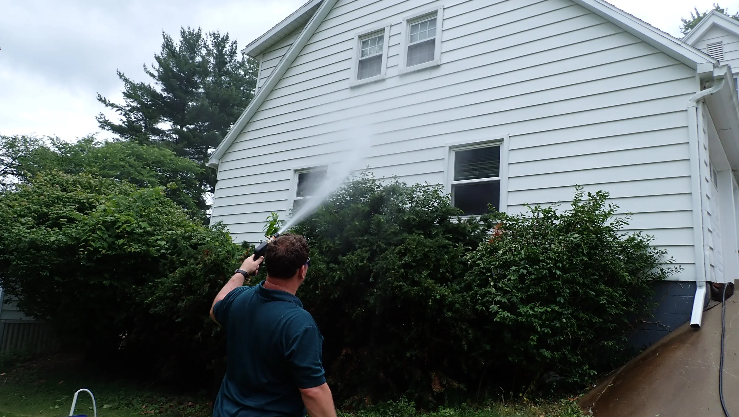 Person standing outside near a white house with an individual using a pressure washer to clean the siding.