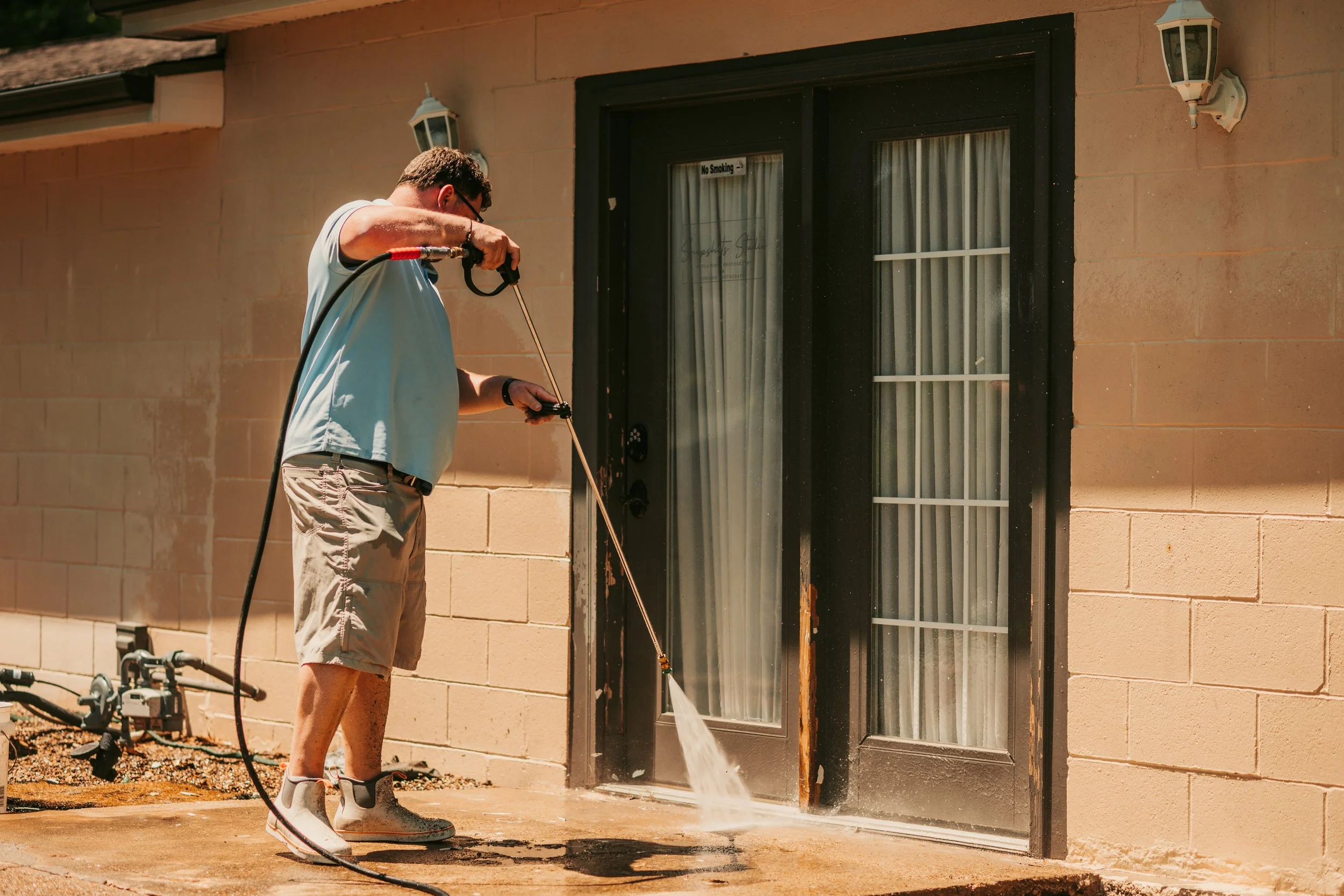 A man wearing beige shorts, a light blue shirt, white sneakers, and glasses is pressure washing the outside of a house at the base of a glass door with a black frame. The house has a beige brick exterior and is illuminated by outdoor lighting fixtures.