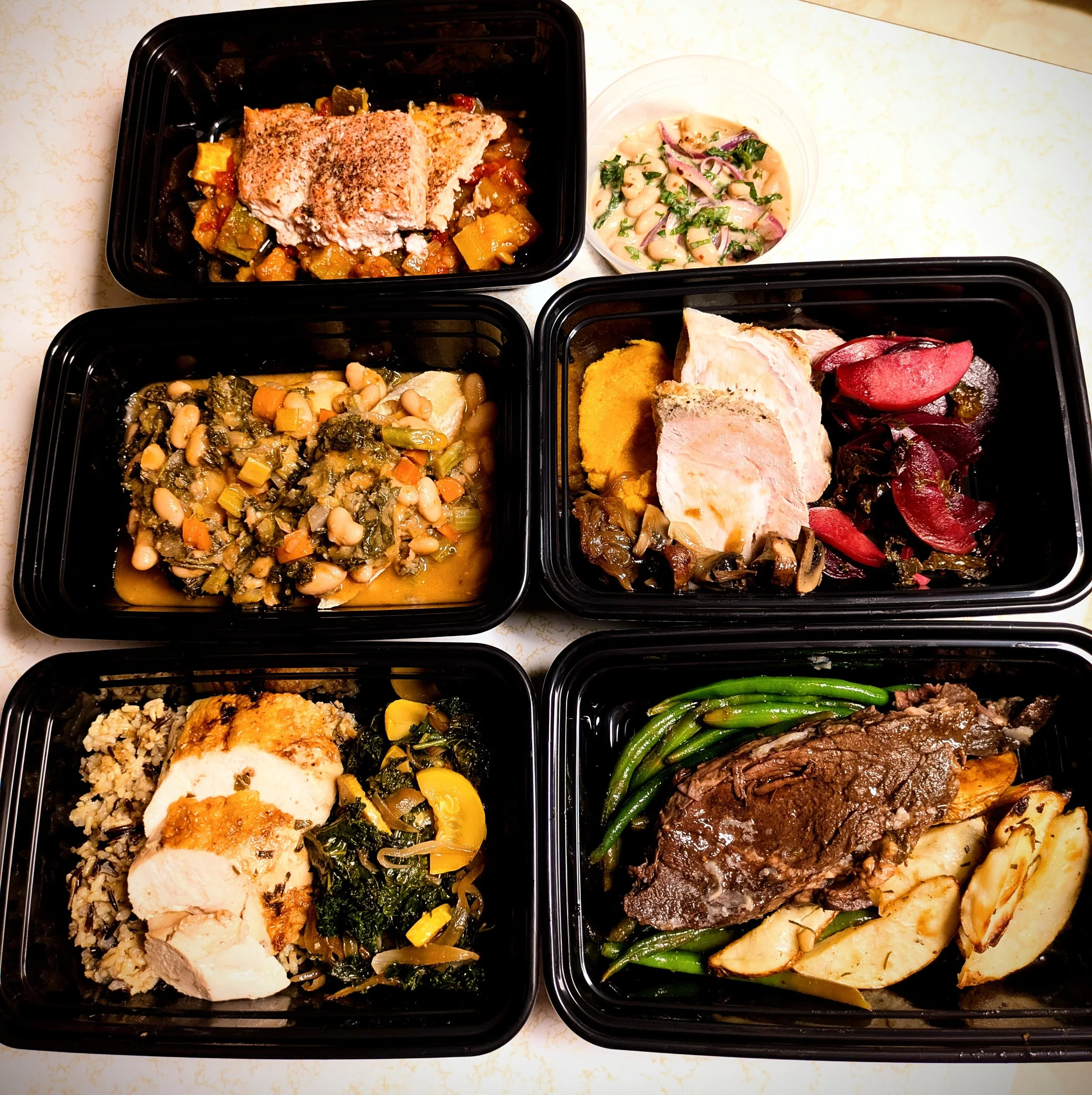 A collection of four meal containers on a white countertop, each filled with various foods. One container has a piece of roast chicken on a bed of rice and mixed vegetables. Another contains a salad with beans, onions, and herbs. A third has cooked greens and sliced yellow squash. The fourth features beef, green beans, and roasted vegetables.
