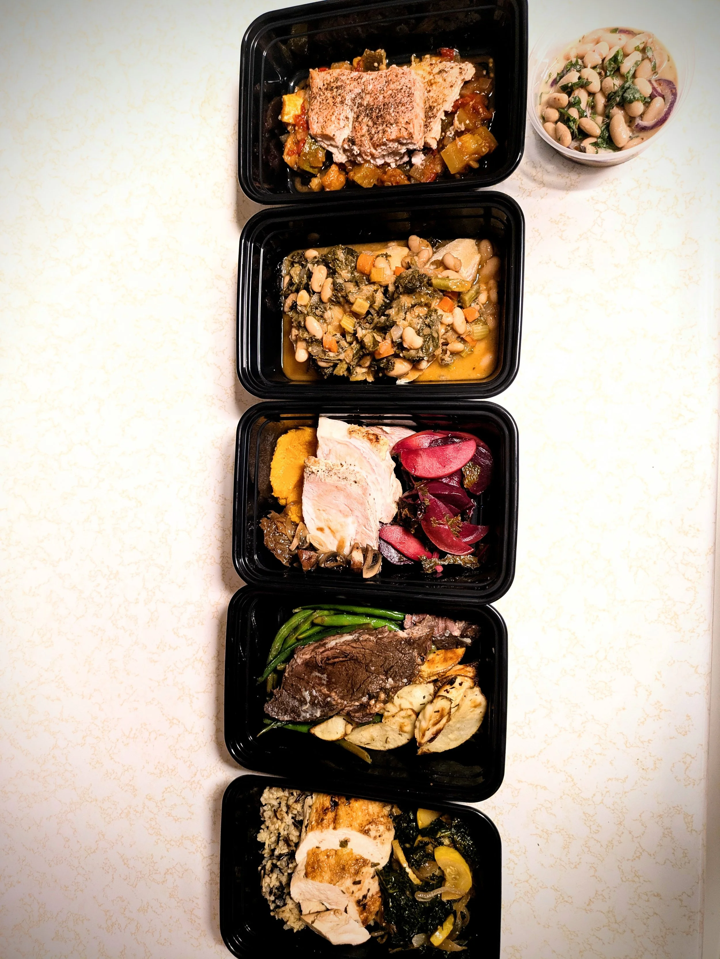 A vertical row of five black meal containers filled with various prepared foods, accompanied by a small plastic cup of bean salad on a light-colored surface.