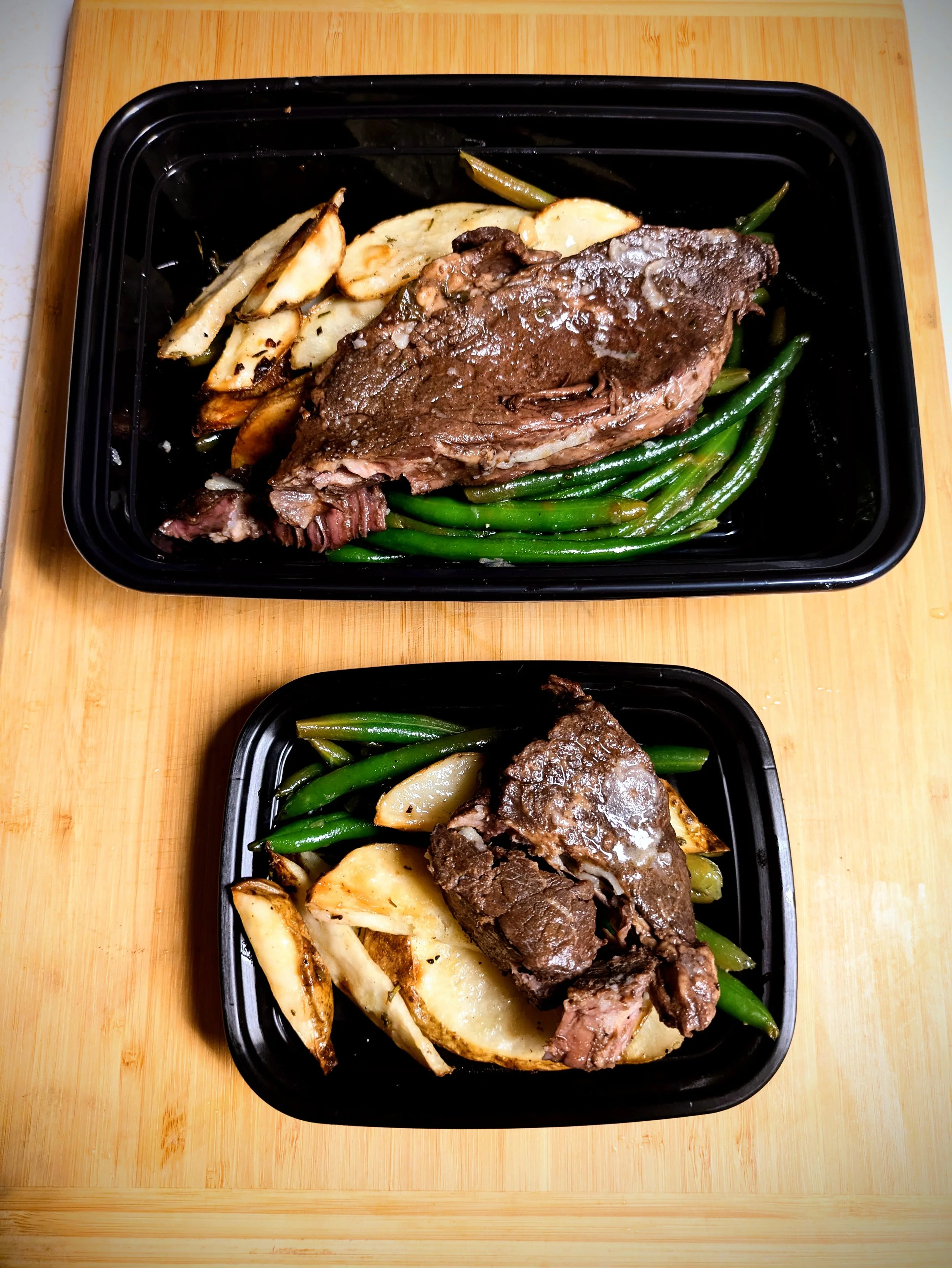 A black meal container with roasted potatoes, green beans, red wine-braised beef, and a large cooked steak, placed on a wooden cutting board.