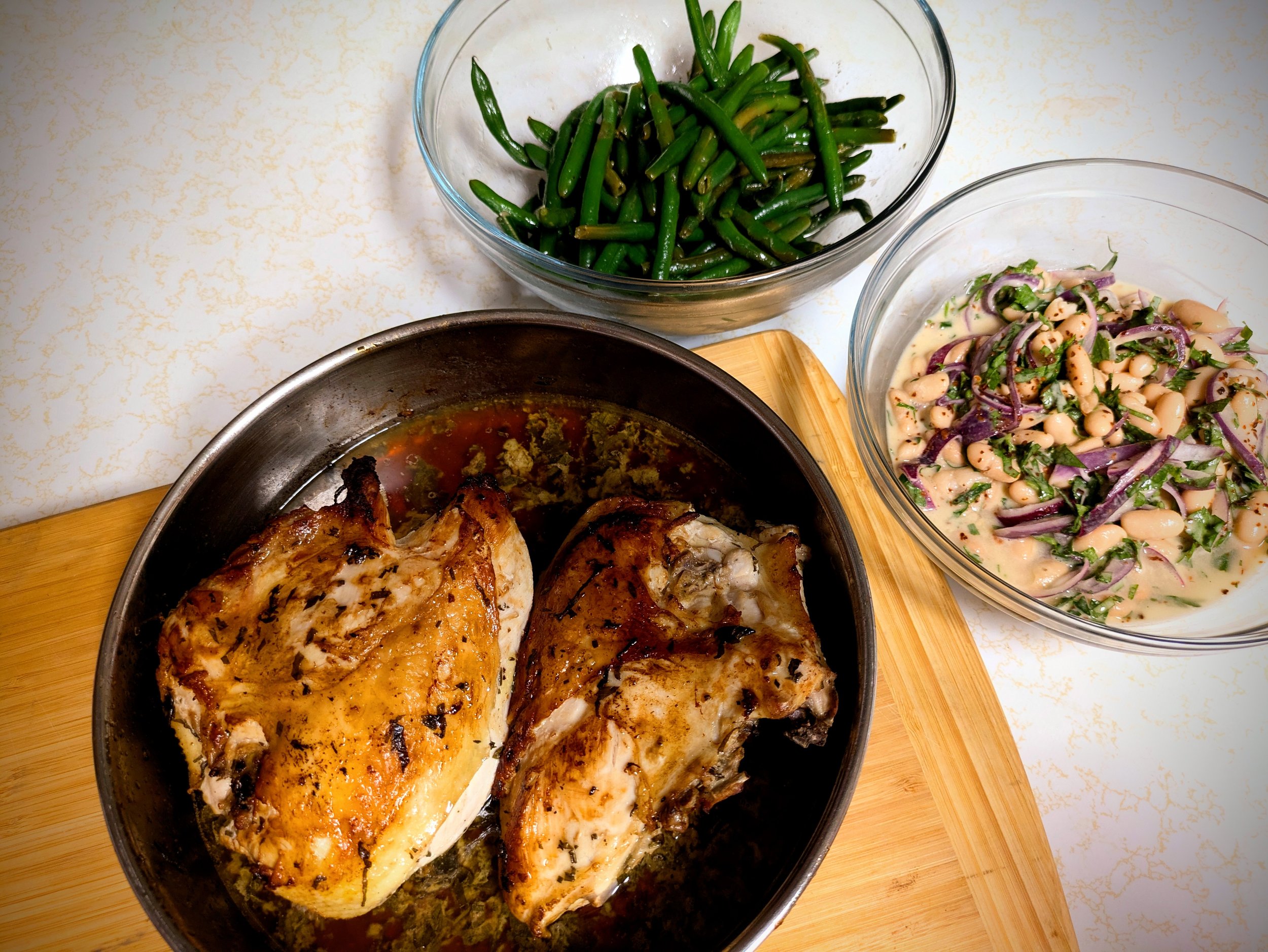 Plate of cooked chicken with a golden brown crust, surrounded by bowls of green beans and bean salad with red onions and herbs.