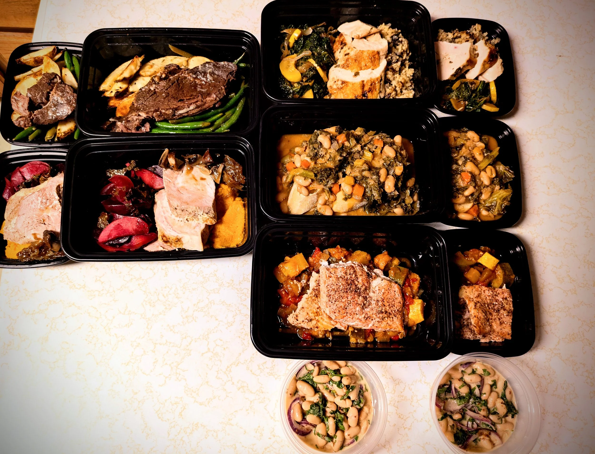 Assorted plastic food containers filled with cooked dishes such as vegetables, meats, beans, and salads arranged on a white countertop.