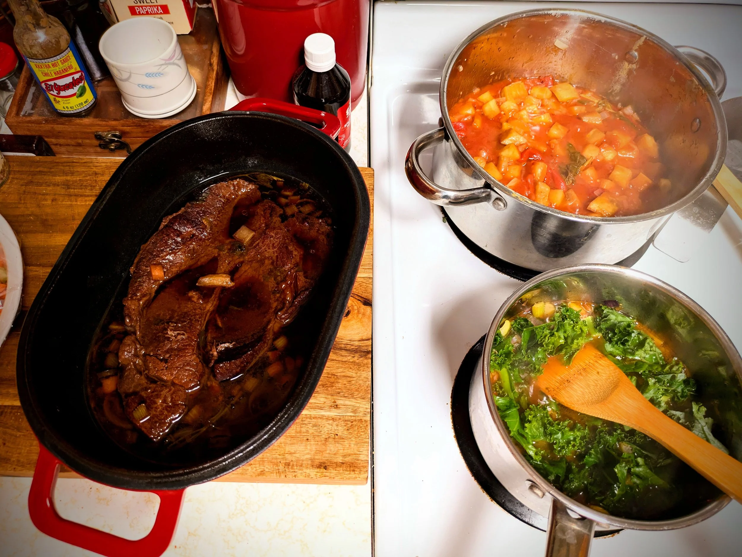 A kitchen countertop with two pots of cooked food, one containing red wine-braised beef and the other containing vegetable stew, surrounded by various condiments, dishes, and utensils.