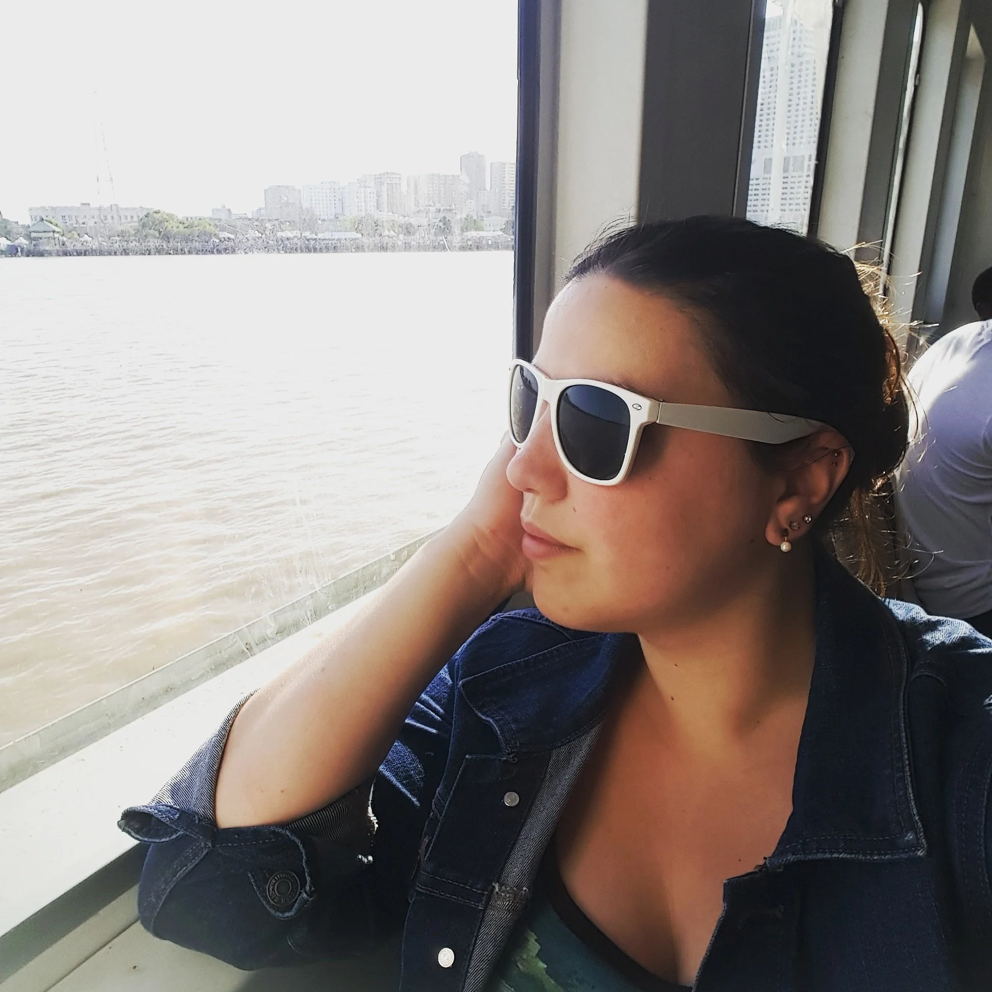A woman wearing white sunglasses and pearl earrings sitting by a window with a view of a cityscape and water.