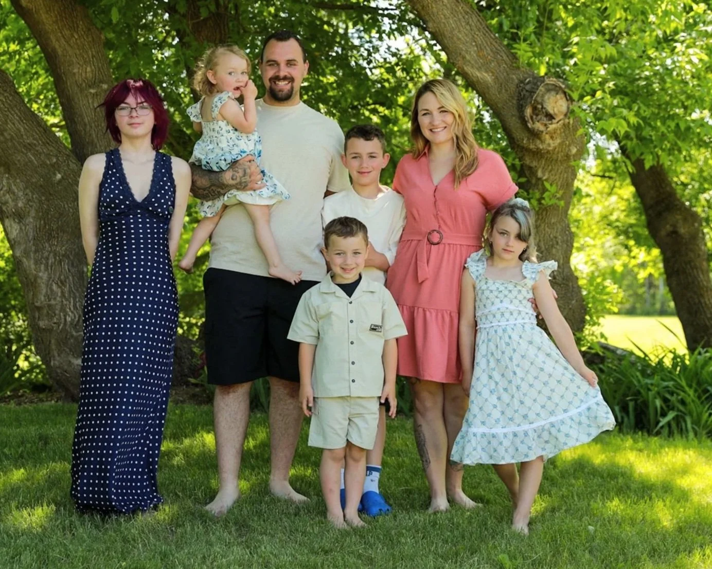 Family of seven standing outdoors under green trees, smiling and posing for a photo on the grass.