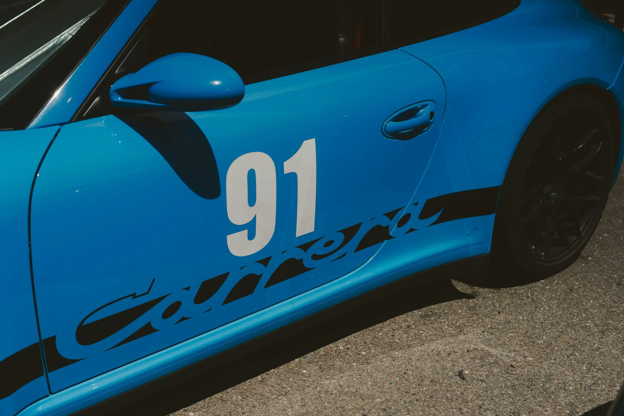 Close-up of a blue sports car with black racing decals and the number 91 on the door, parked on a gravel road.