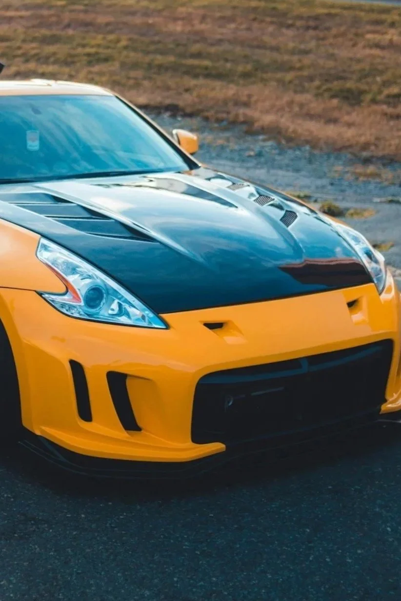 A yellow sports car with a black hood, parked on a road with grass and dirt in the background.