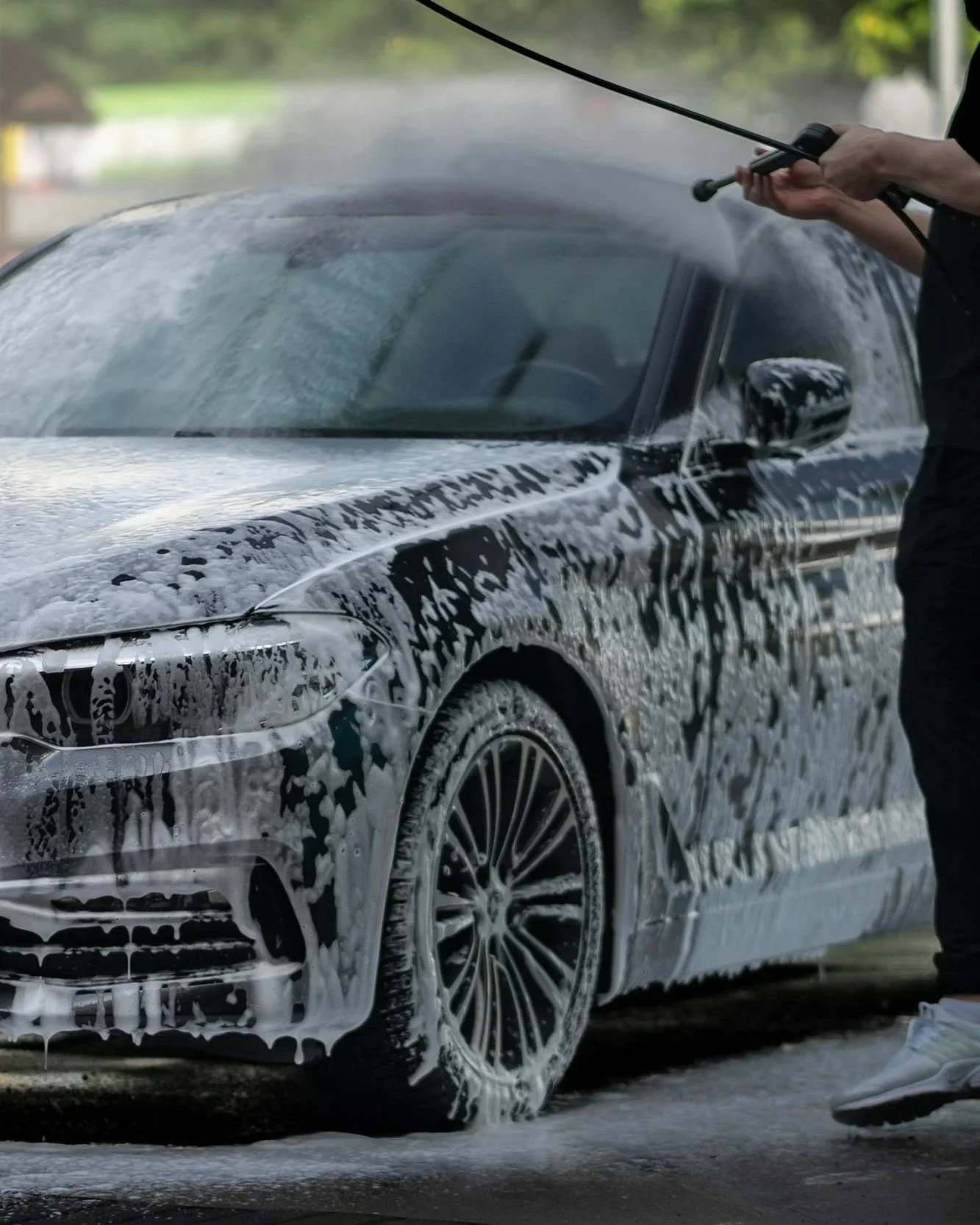 A person washing a black car with soap and water, creating foam on the car's surface.