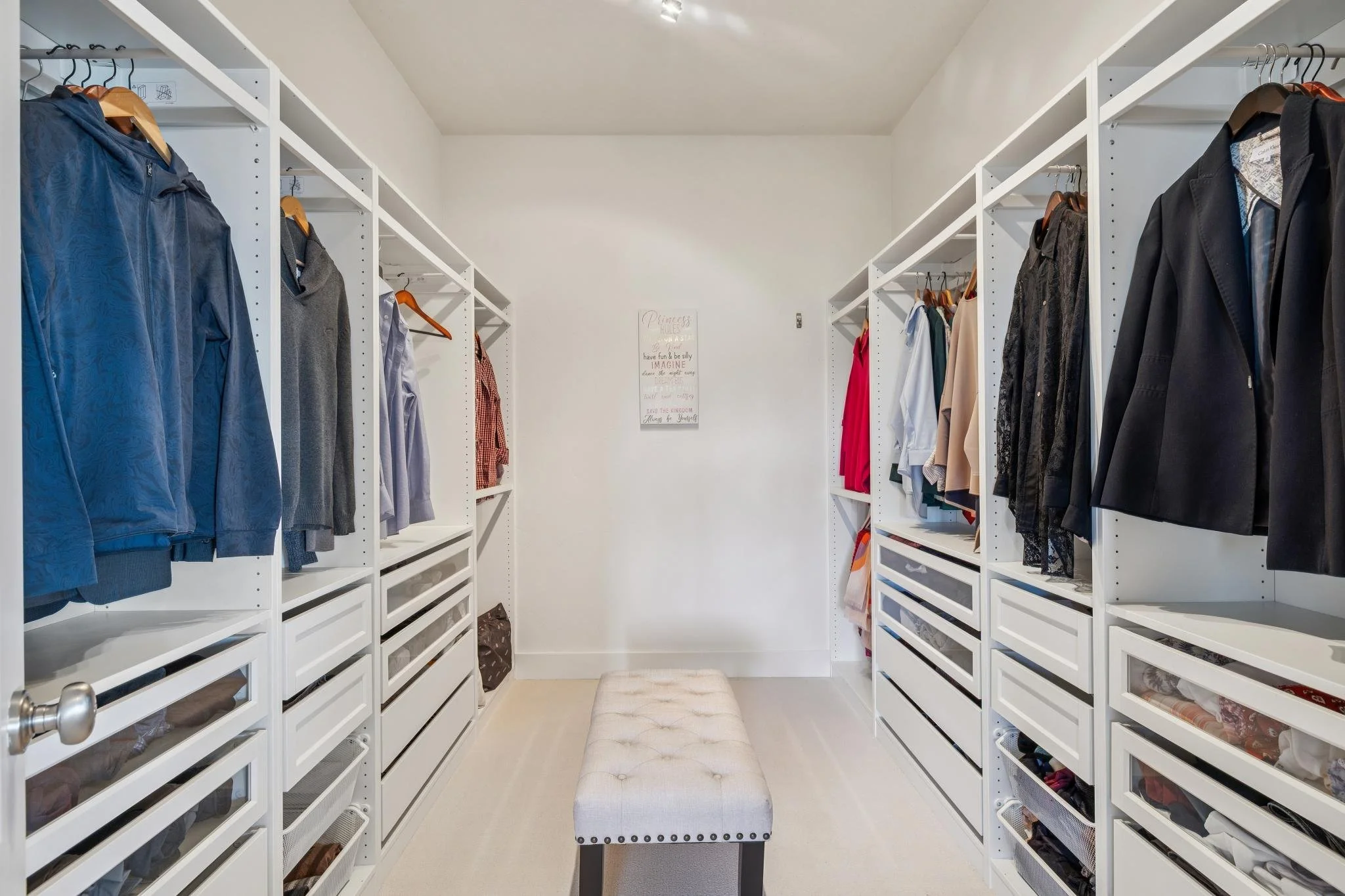 Walk-in closet with white shelving units on both sides filled with clothing and accessories, small bench in the center, and a framed quote on the back wall.