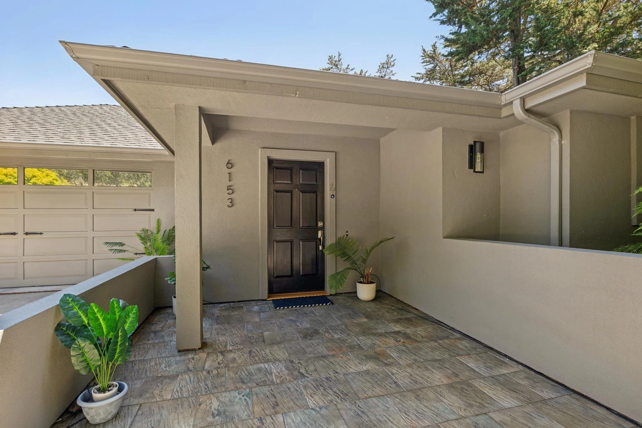Front porch of a house with potted plants, a dark brown door, a white garage door in the background, and a light-colored wall with house numbers 6153, under a blue sky.